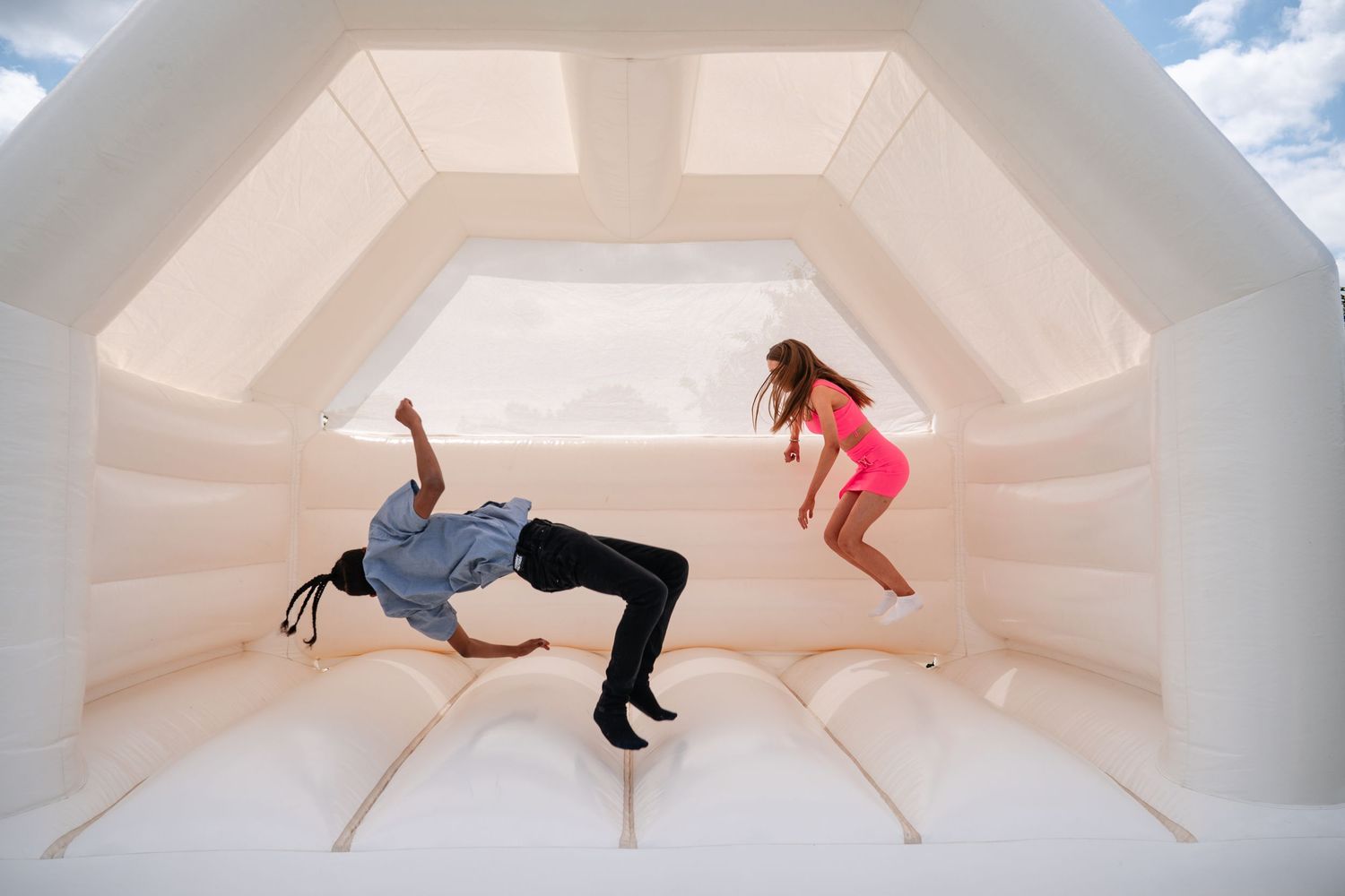 Two people jumping in a white bounce house, one in pink outfit and one in dark clothing, mid-air against white walls.