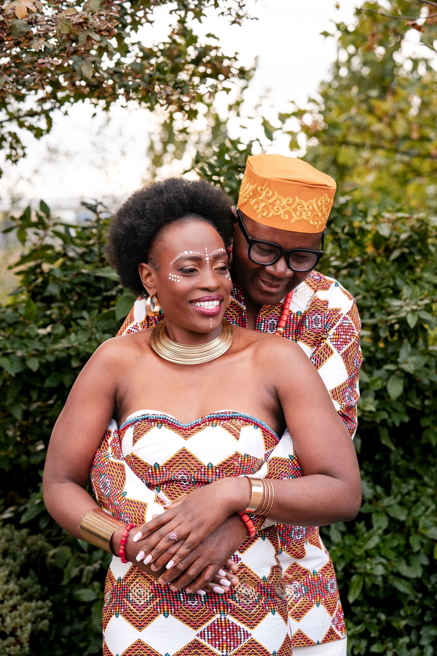 Couple in vibrant African traditional attire embracing outdoors, woman wearing beaded dress and gold jewelry, man in matching cap.