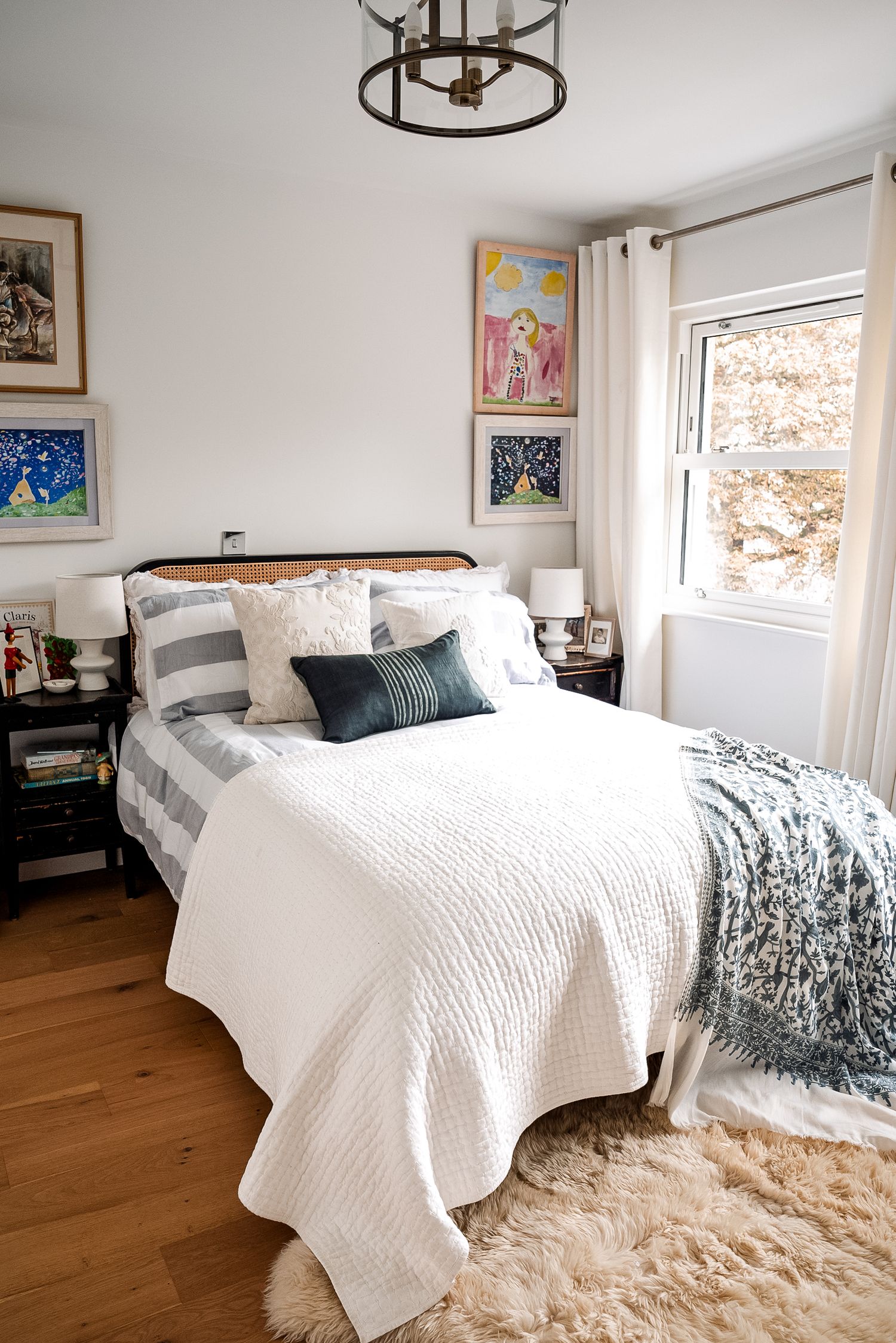 Cozy bedroom with white bedding, dark throw pillows, gallery wall art, shag rug, and natural light from a corner window.