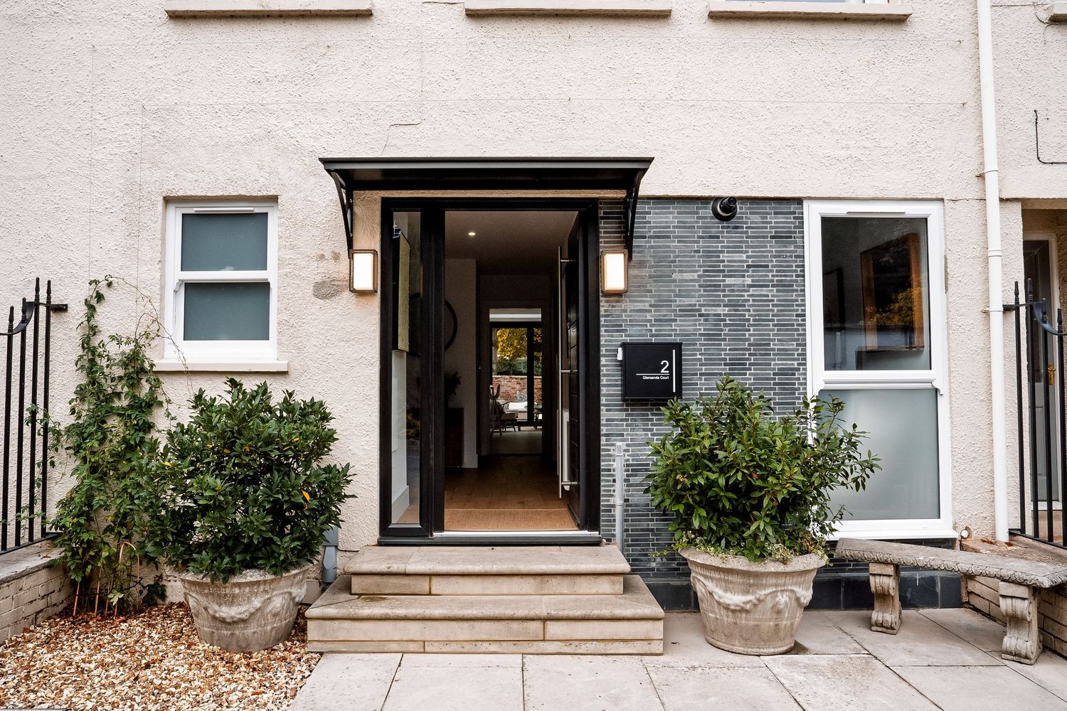 Modern black front door entrance with stone steps, potted shrubs, and elegant wall lights on a white rendered facade.