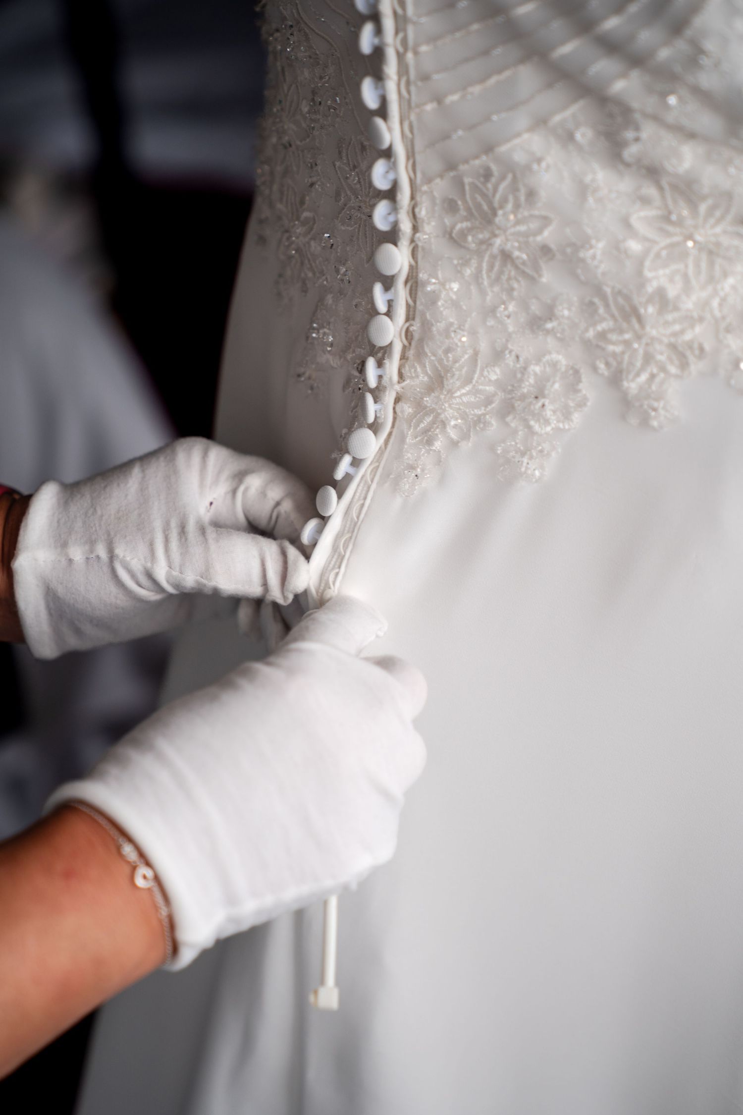 Gloved hands carefully fastening delicate pearl buttons on an elegant white lace wedding dress.