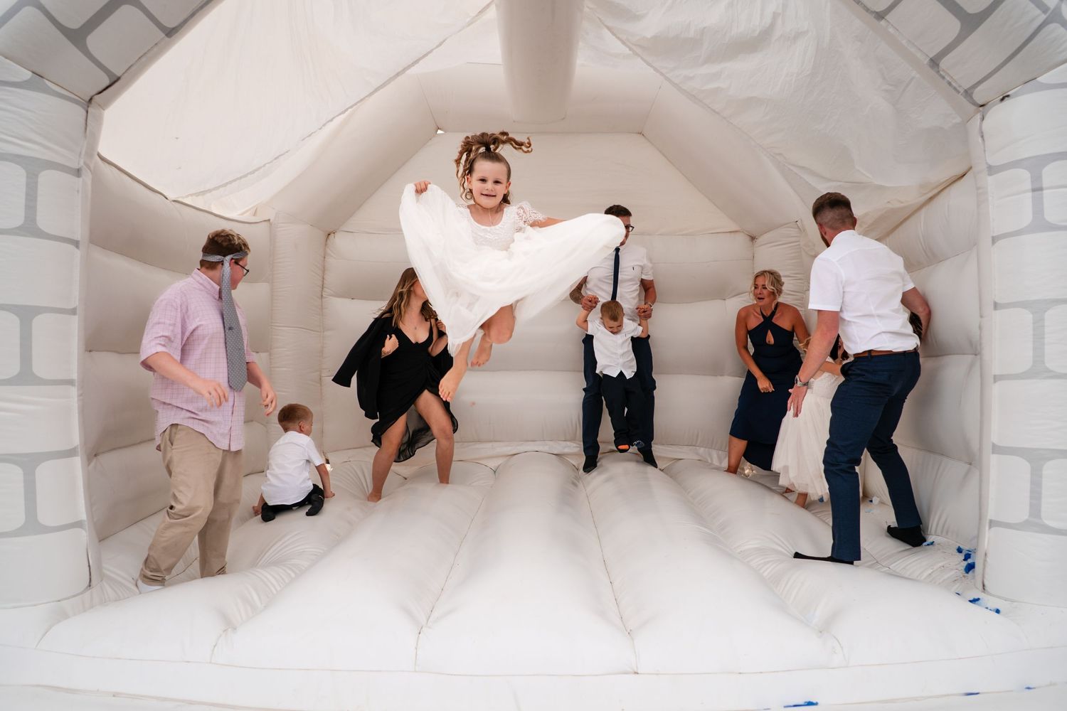 Bride in white wedding dress jumping joyfully in a white bounce house with guests at a wedding reception.