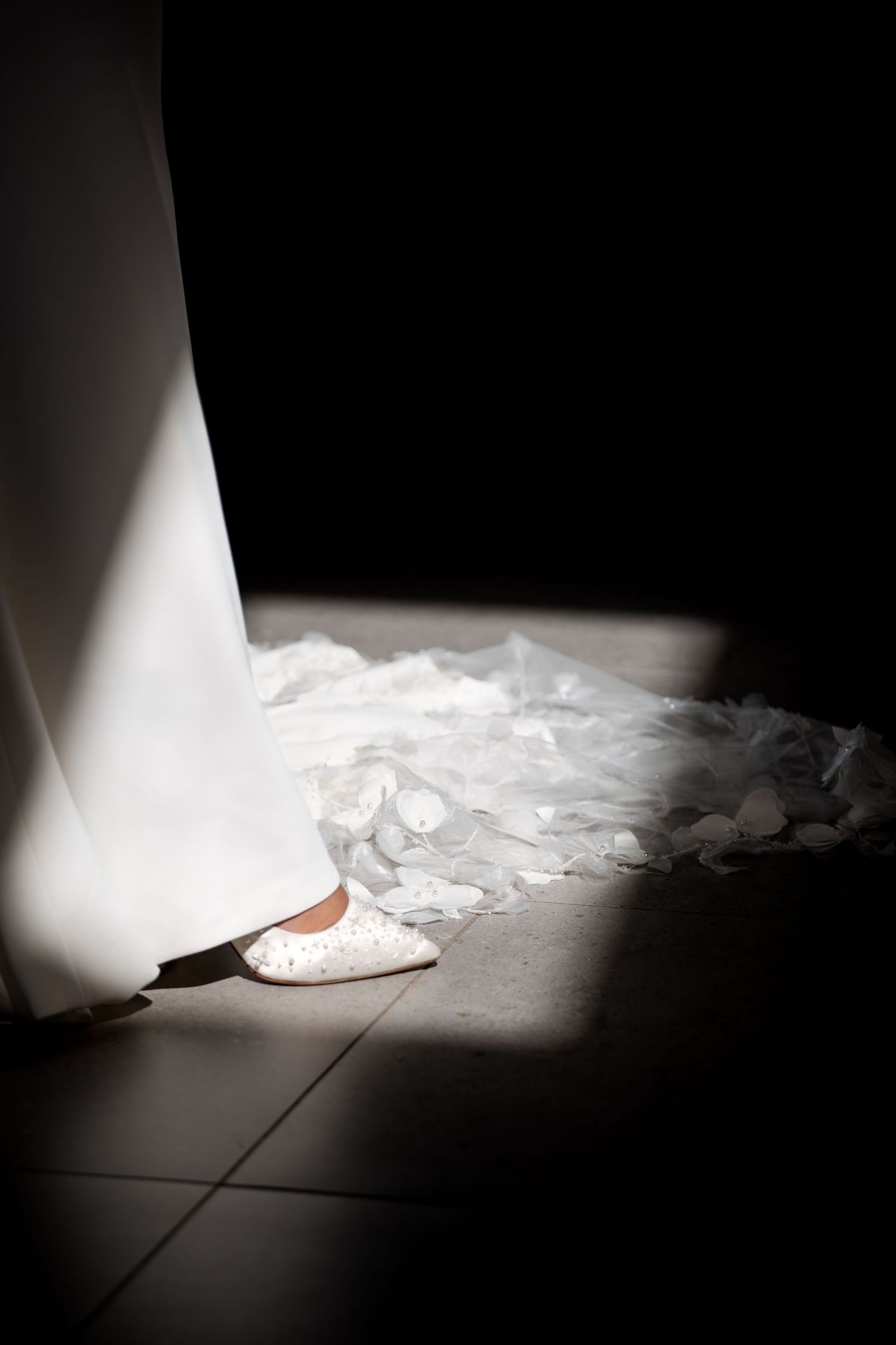 A bride's white lace wedding shoe peeks beneath her gown and veil in a dramatic beam of light on a tiled floor.