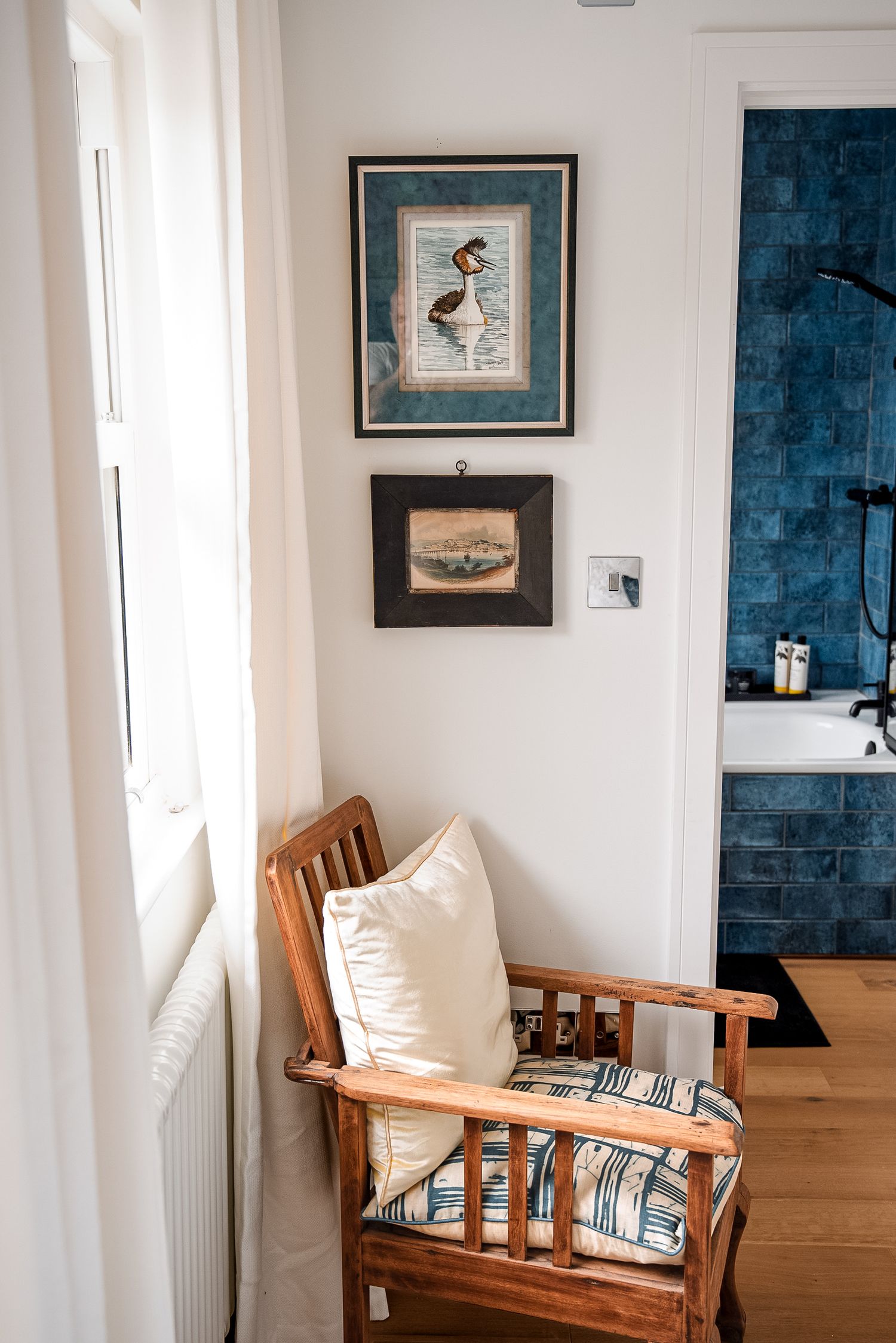 Wooden armchair with white cushion beside a window, two framed bird prints on white wall, blue tiled bathroom visible.
