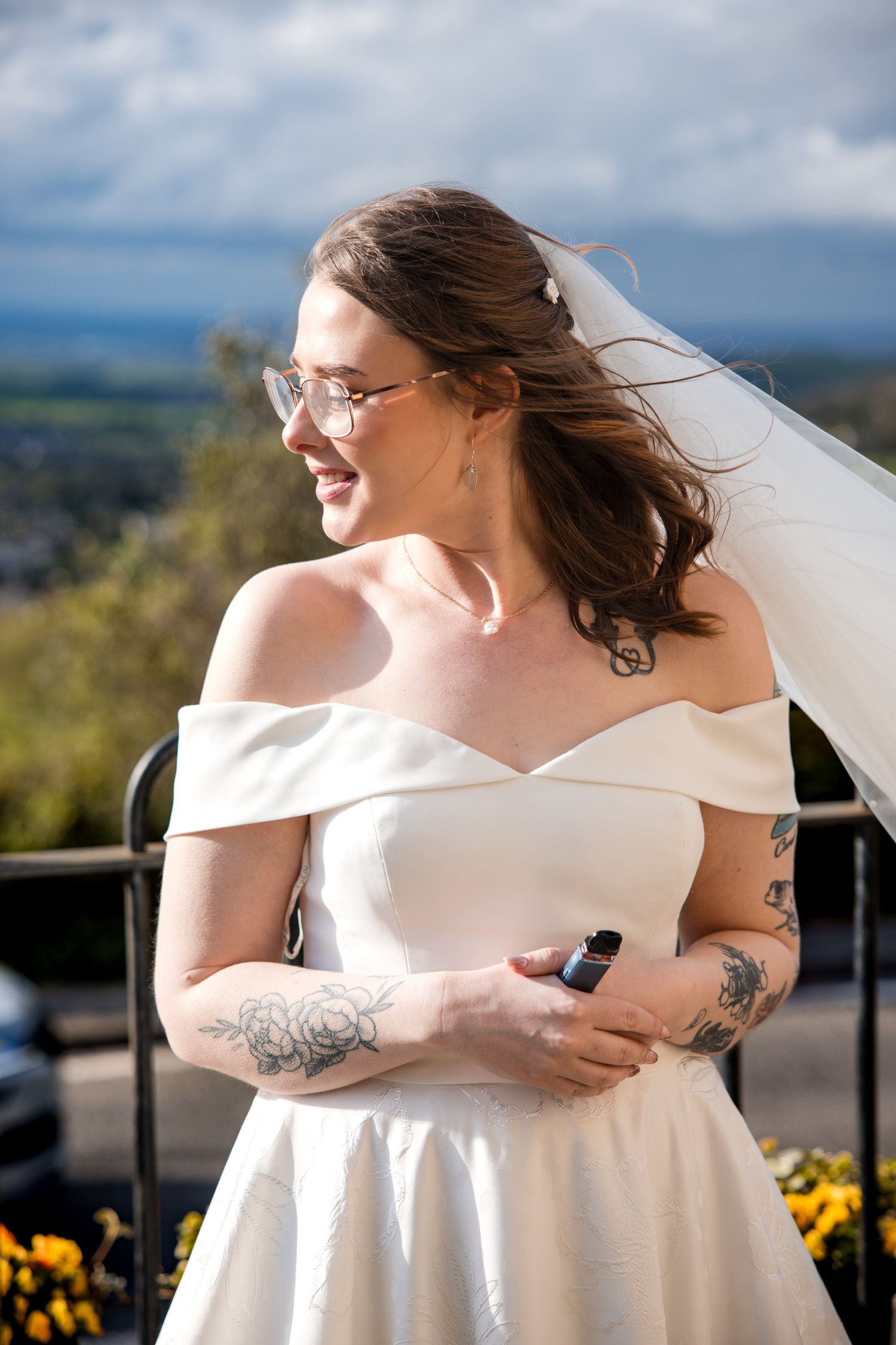 Smiling bride in off-shoulder white gown and veil, wearing glasses and tattoos, standing outdoors on a sunny day.
