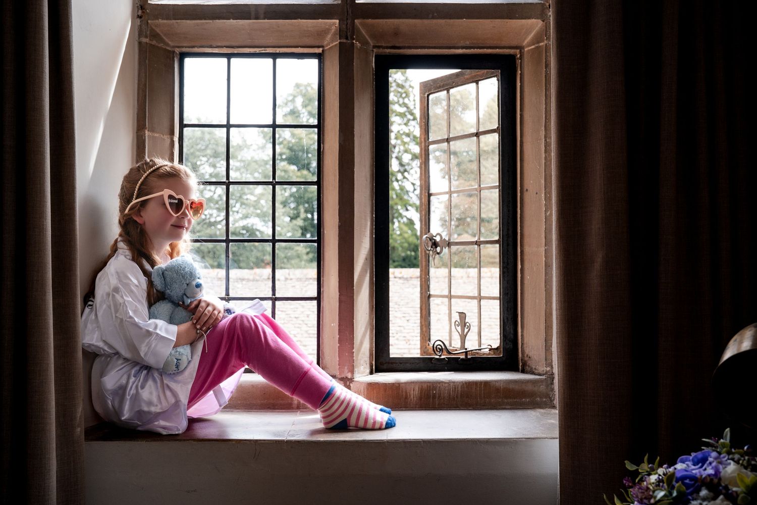 Young girl in pink pants and heart glasses sits on a stone windowsill hugging a stuffed toy, gazing outside.
