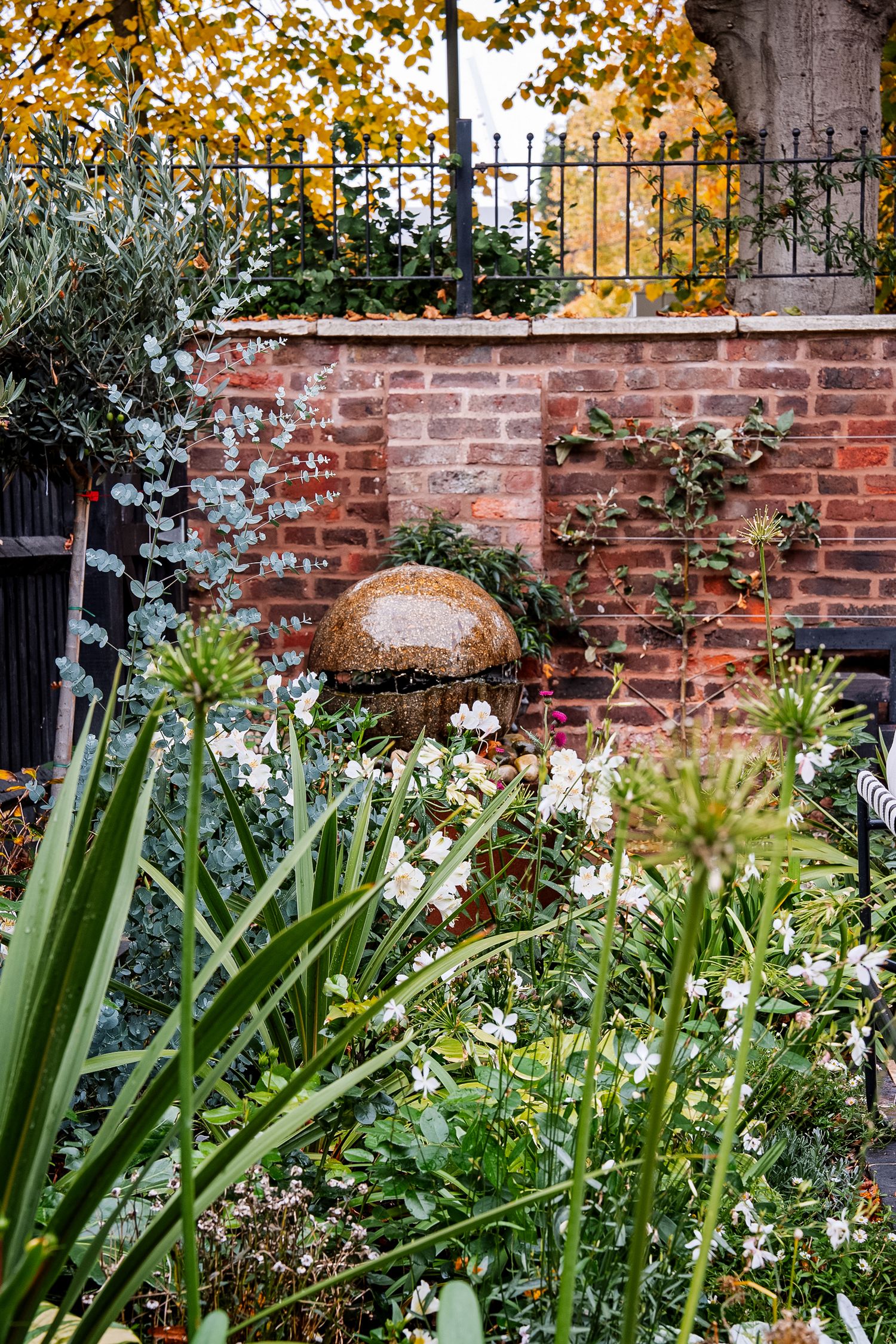 Lush garden with white flowers, green plants, and a decorative stone sphere against a rustic red brick wall with iron fence.