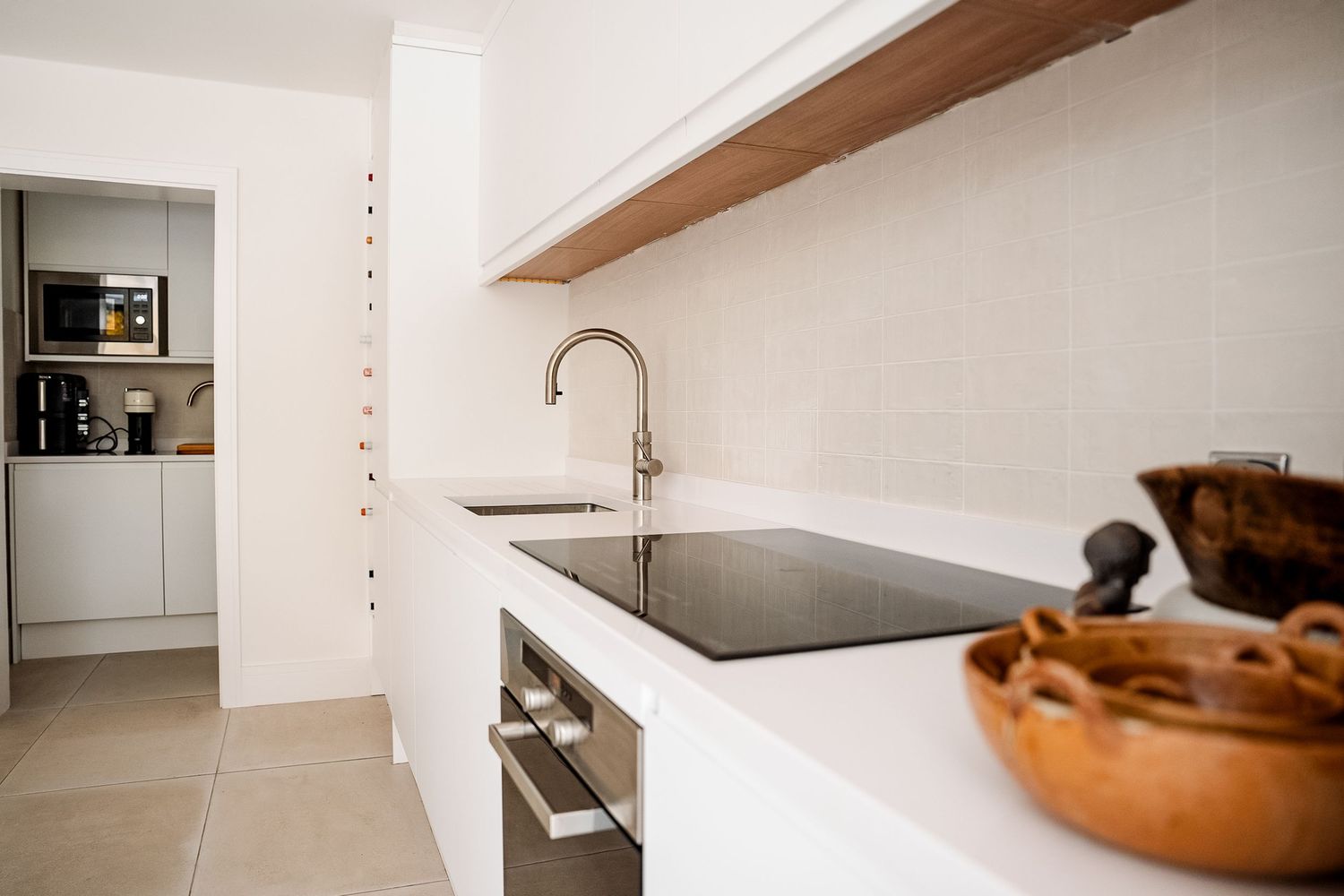 Modern white kitchen with black sink, chrome faucet, wooden shelf, and built-in appliances in a minimalist design.