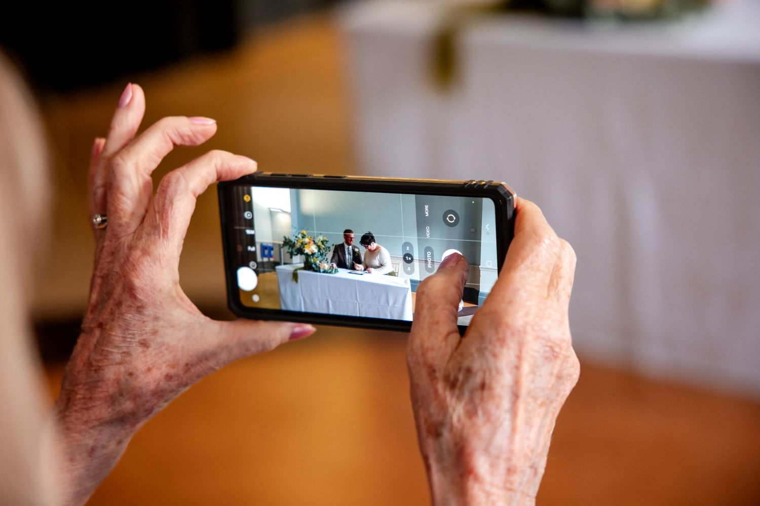 Elderly person holding smartphone horizontally, photographing a group of people seated at a table during an event.