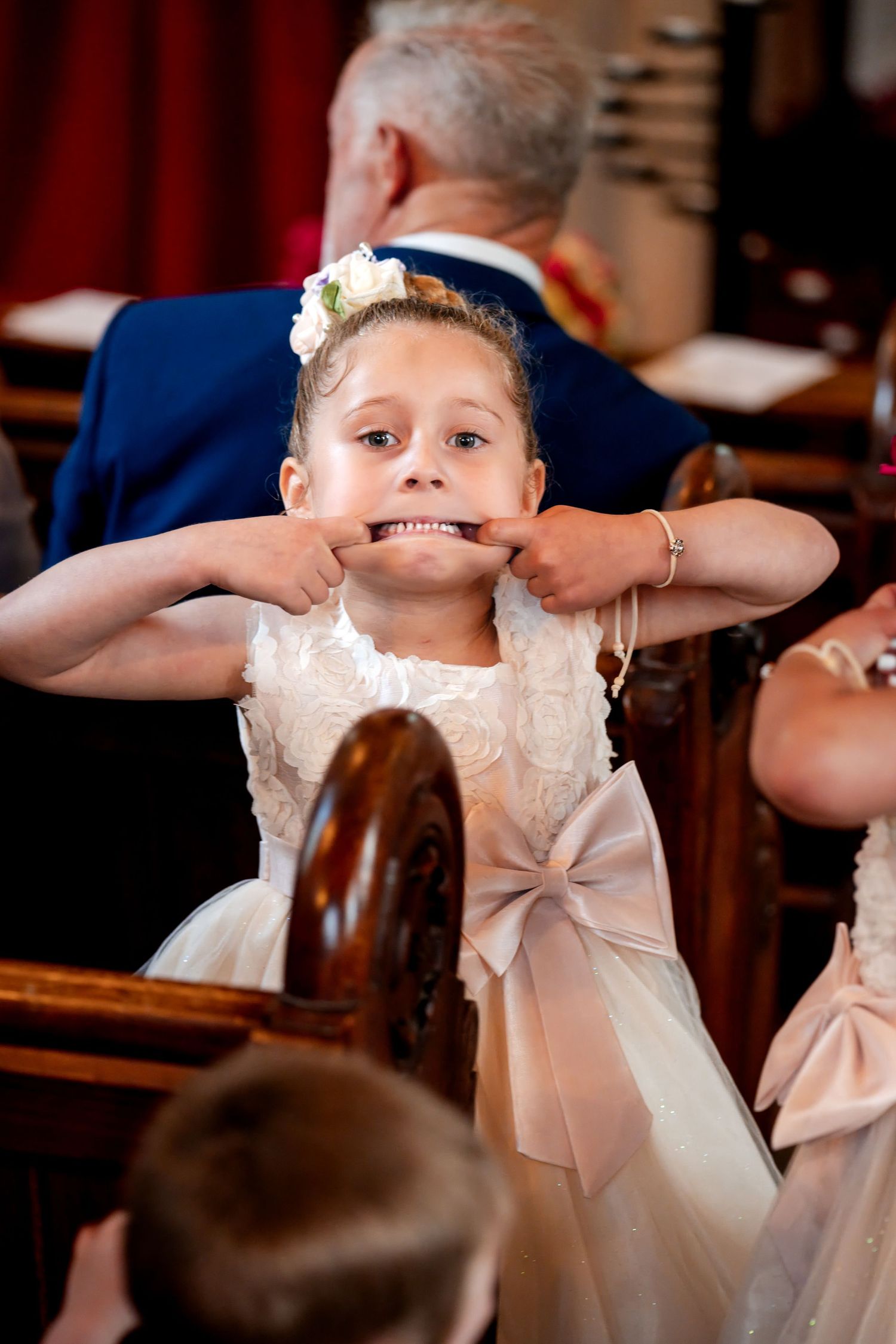Flower girl in white dress making a funny face by stretching her mouth with fingers during a wedding ceremony.
