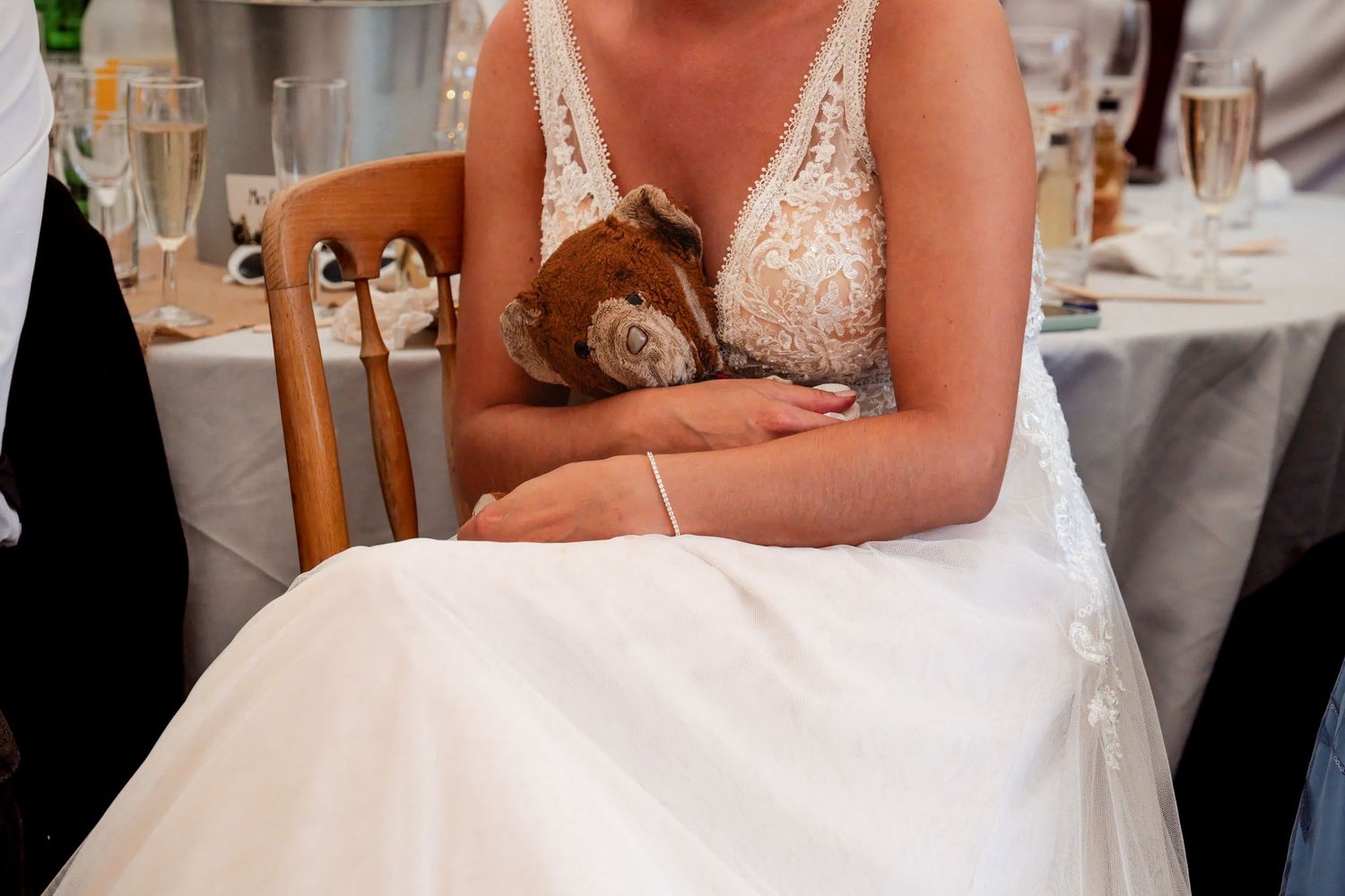 Bride in white lace wedding dress holding a brown teddy bear while seated at a reception table.