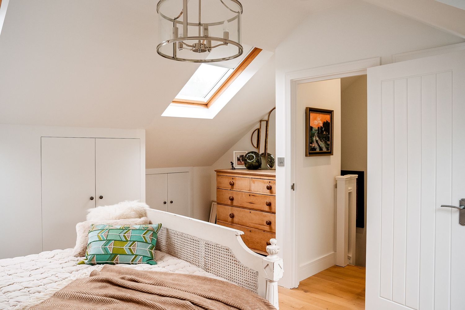 Bright attic bedroom with skylight, wicker bed, pine dresser, white wardrobes, and chandelier on light wood floors.