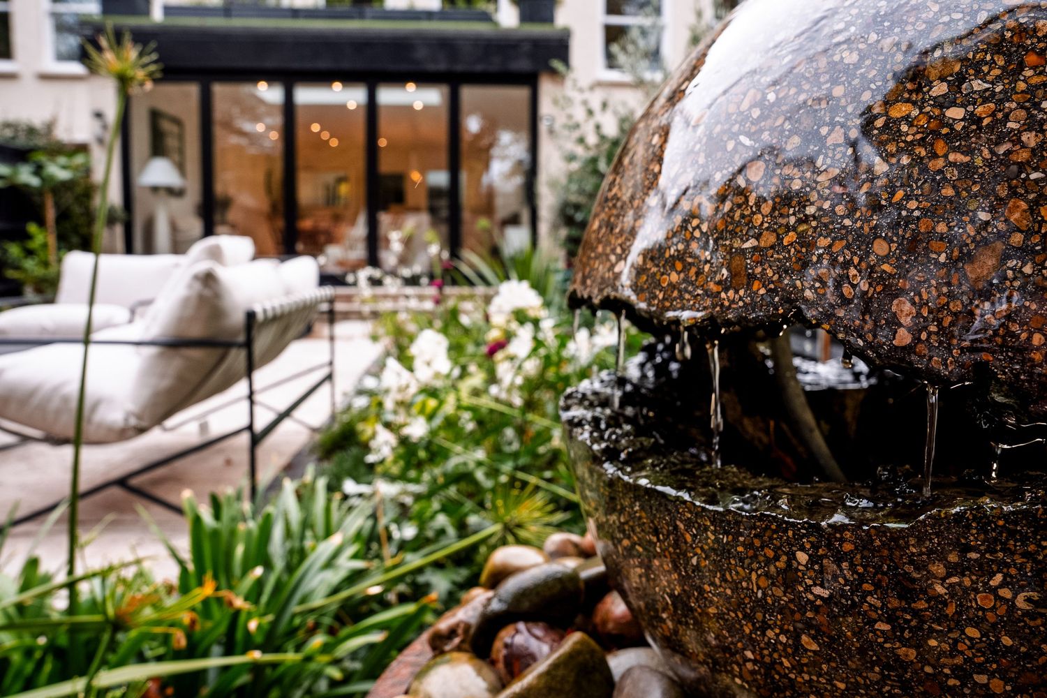 A decorative granite sphere water fountain surrounded by lush greenery and white flowers in an elegant outdoor garden.