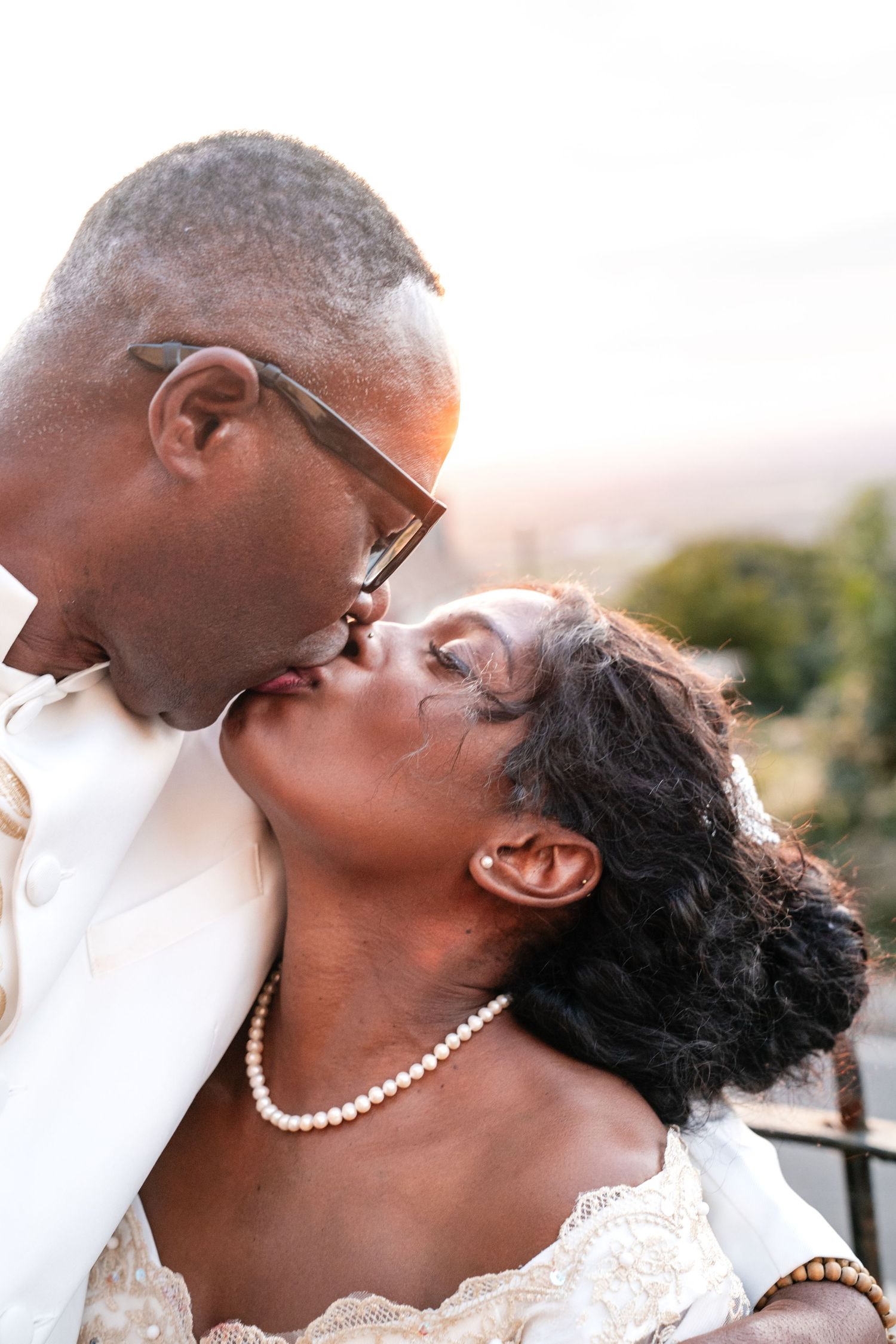 A Black bride and groom share a romantic kiss outdoors, dressed in elegant white wedding attire with pearl jewelry.