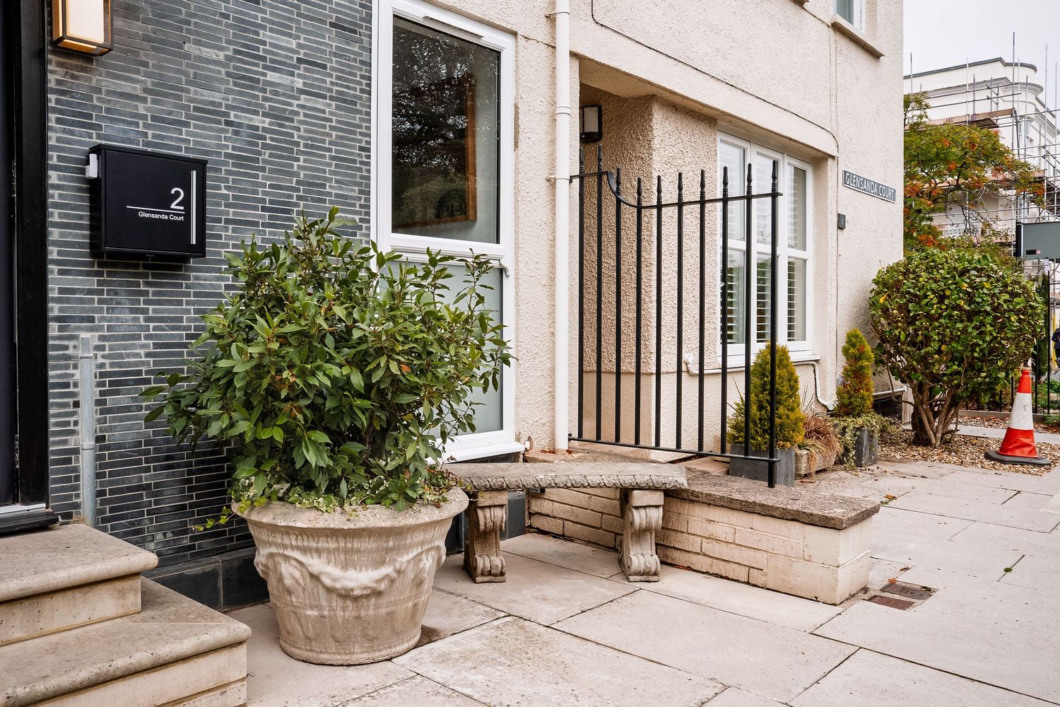 Elegant building entrance with stone bench, large potted shrub, and wrought iron gate on a paved courtyard.