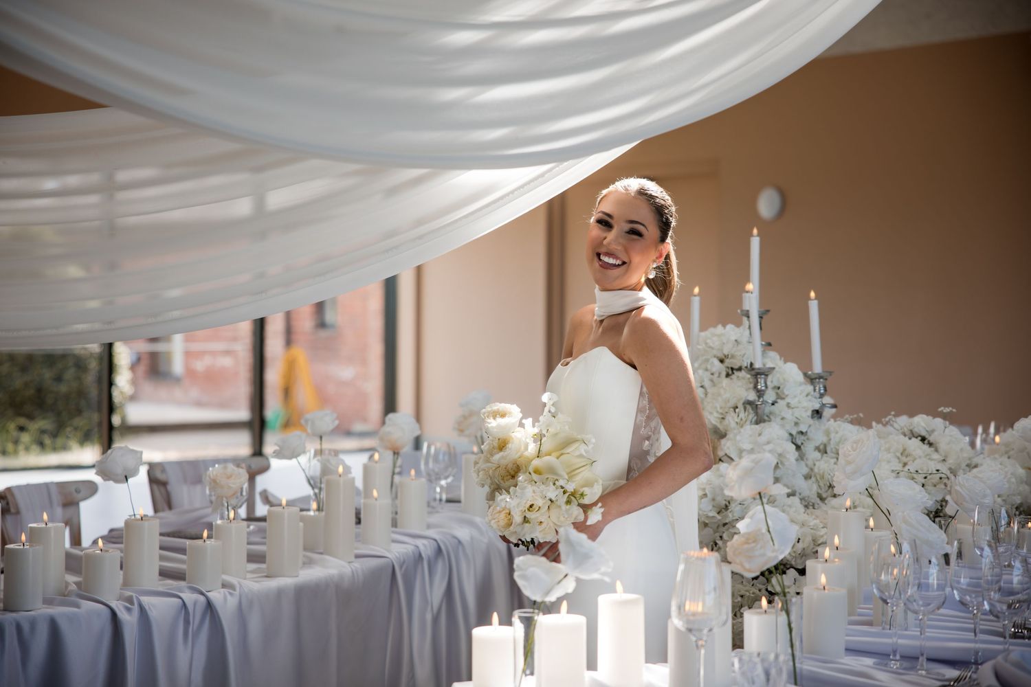 Smiling bride in white gown holding bouquet stands beside elegant white candle and floral wedding reception table decor.
