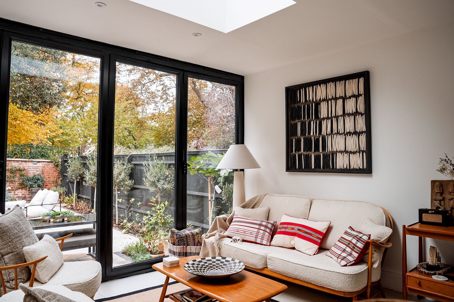 Bright living room with white sofa, red pillows, wood coffee table, and large black-framed glass doors opening to autumn garden.