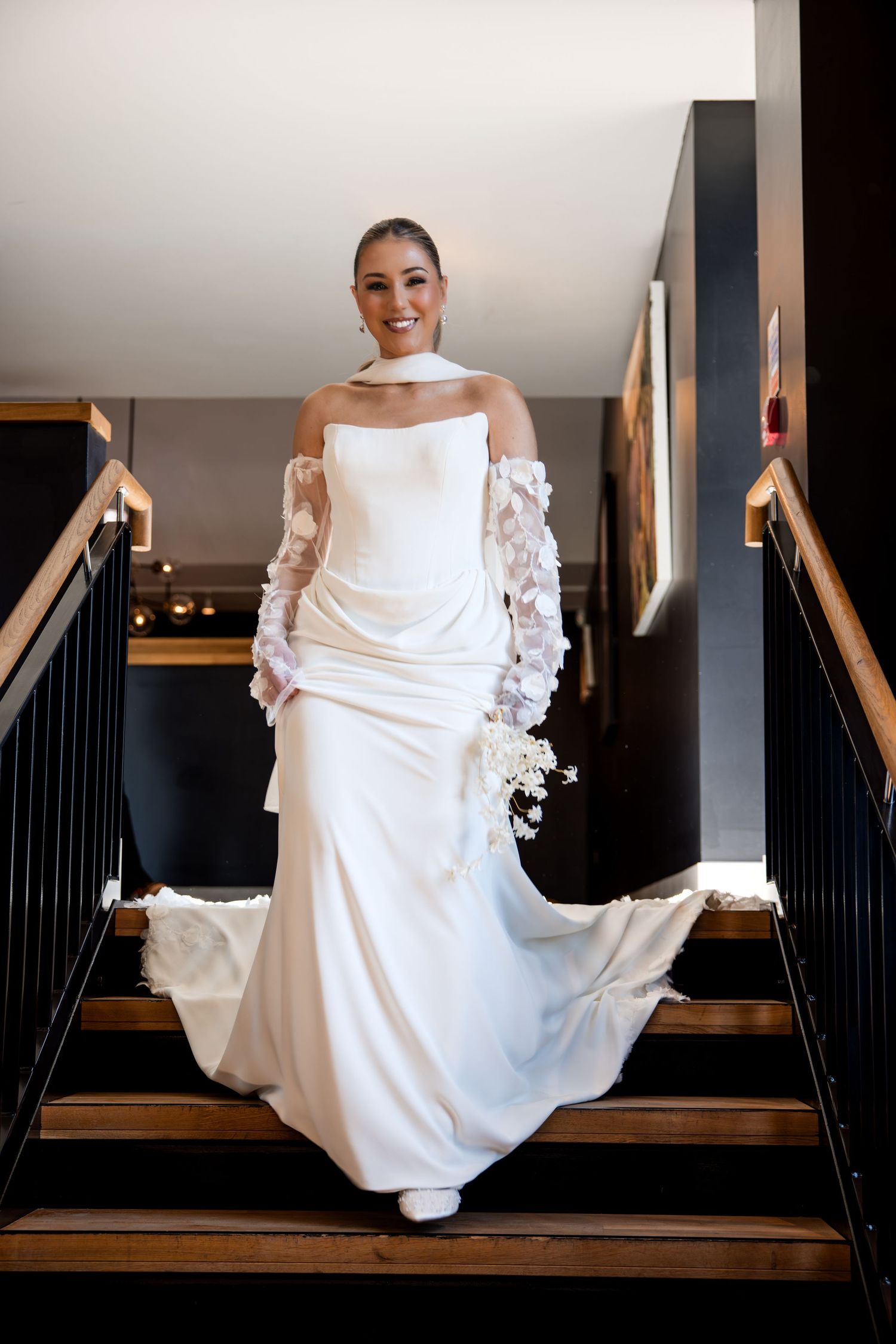 Bride in white strapless gown with lace gloves holding bouquet descending elegant staircase.