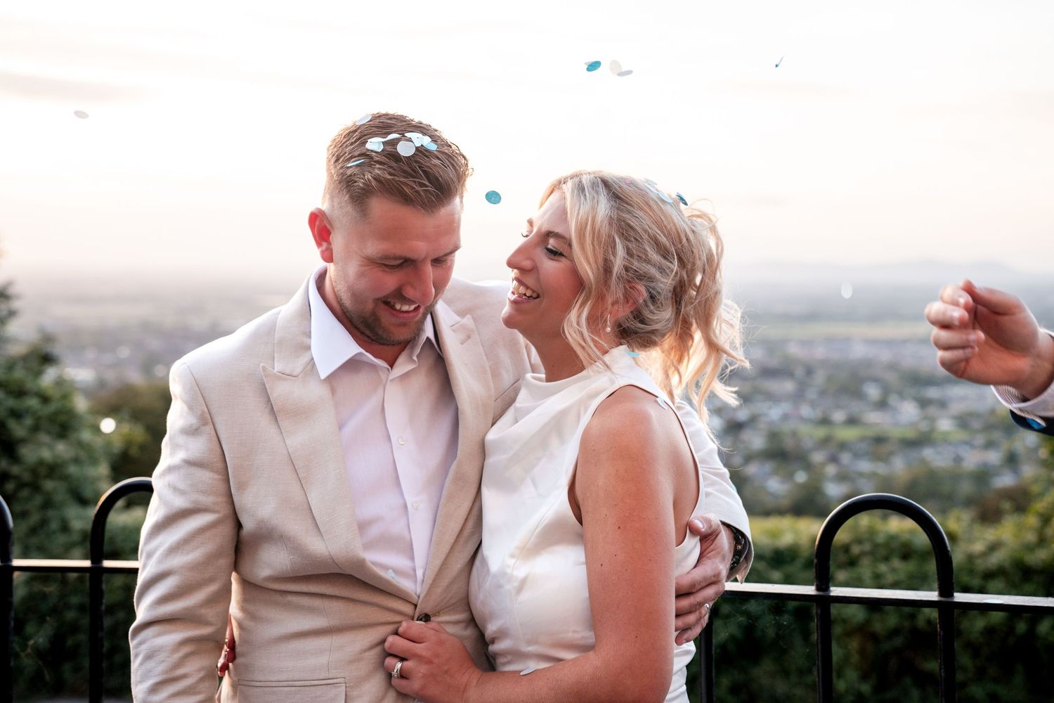 Smiling newlywed couple in wedding attire embracing outdoors with confetti and scenic city views at sunset.