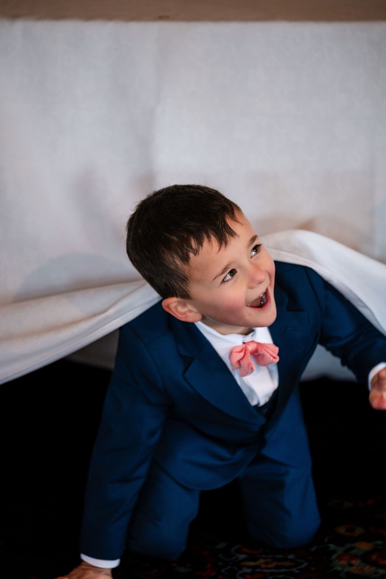 Young boy in navy suit and pink bow tie crawling under white fabric, smiling joyfully at a wedding.