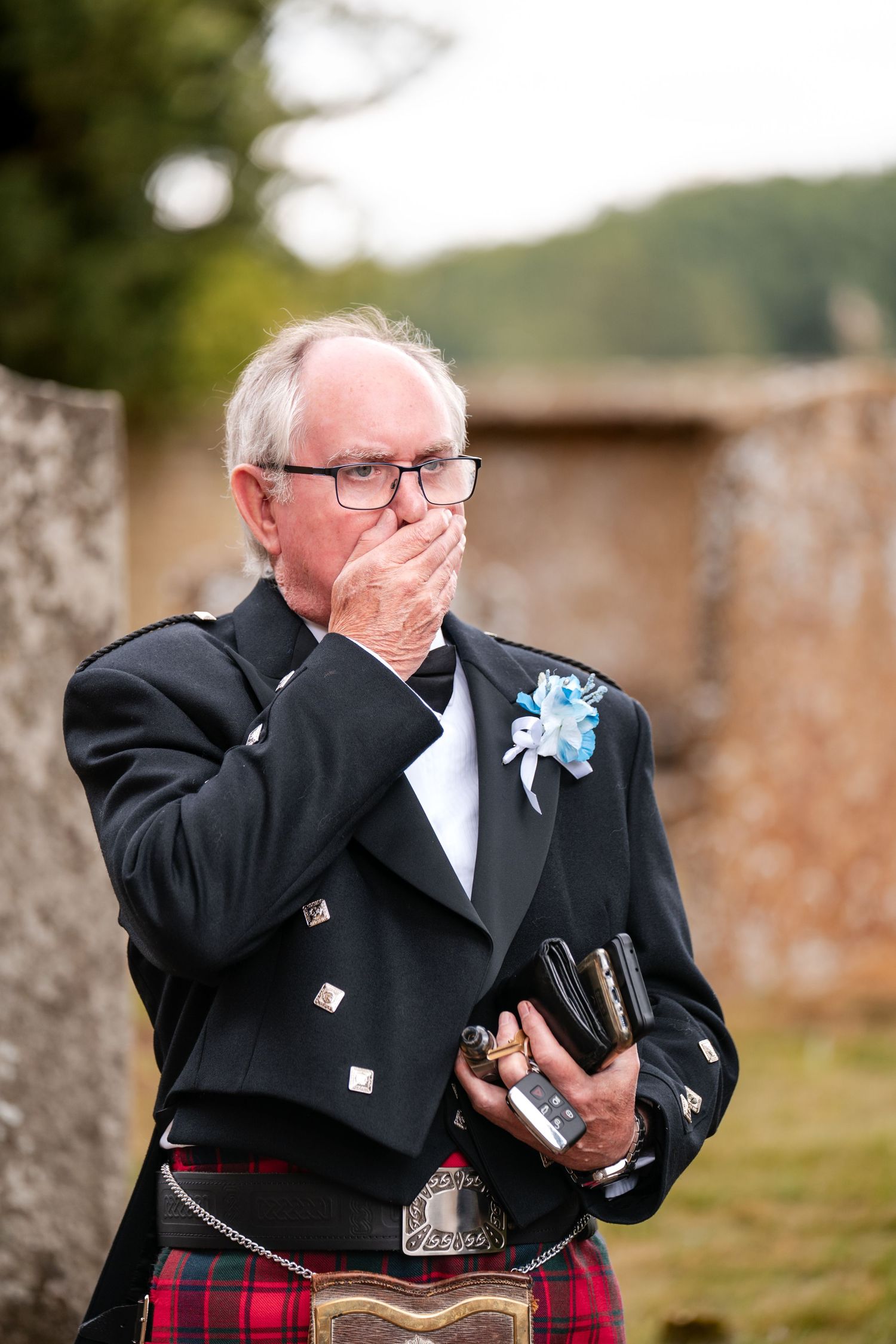 Elderly man in Scottish kilt and black jacket with blue boutonniere covers mouth emotionally outdoors.