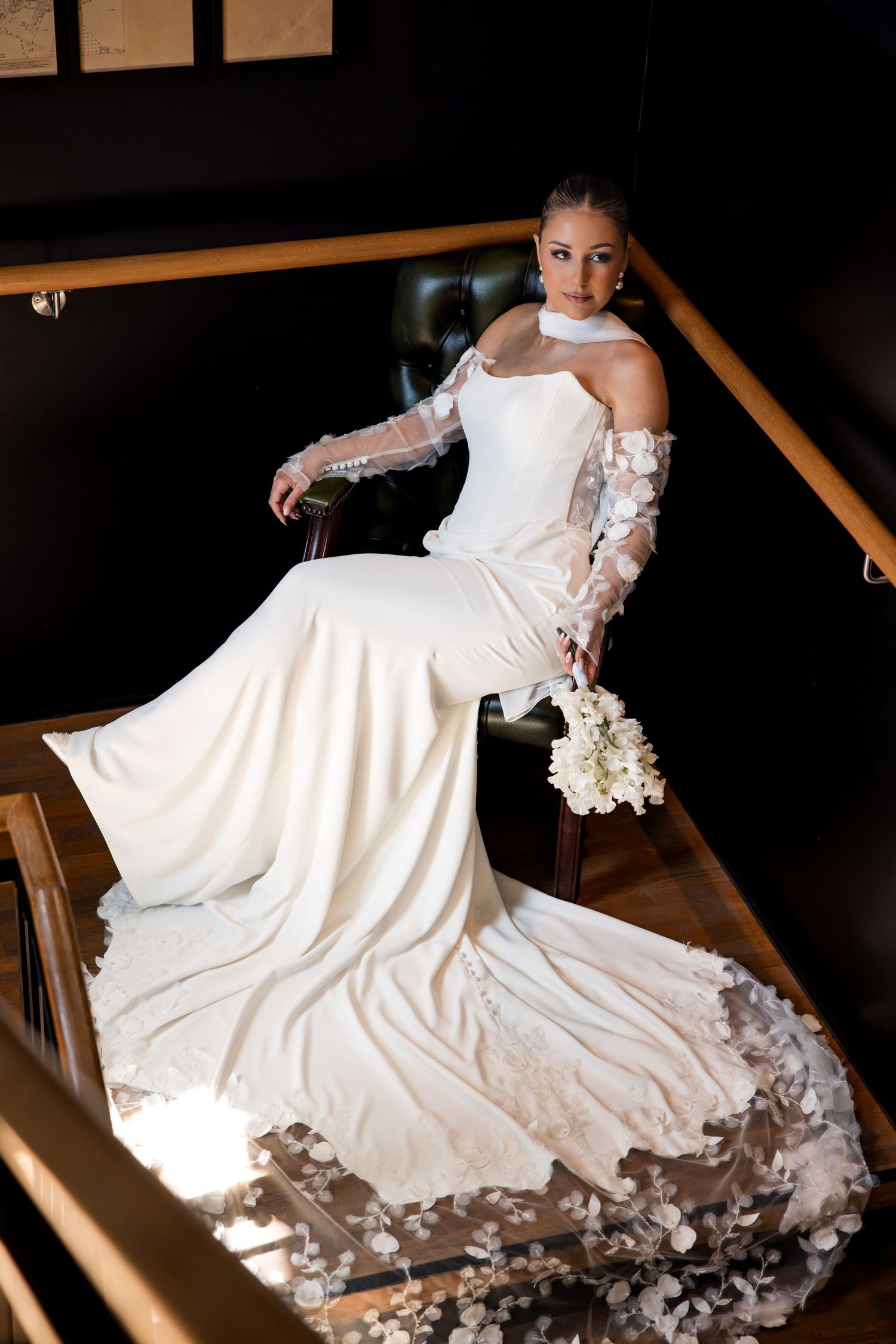 Elegant bride in white gown with floral sleeves sits on stairs holding bouquet, dress train spread beautifully.