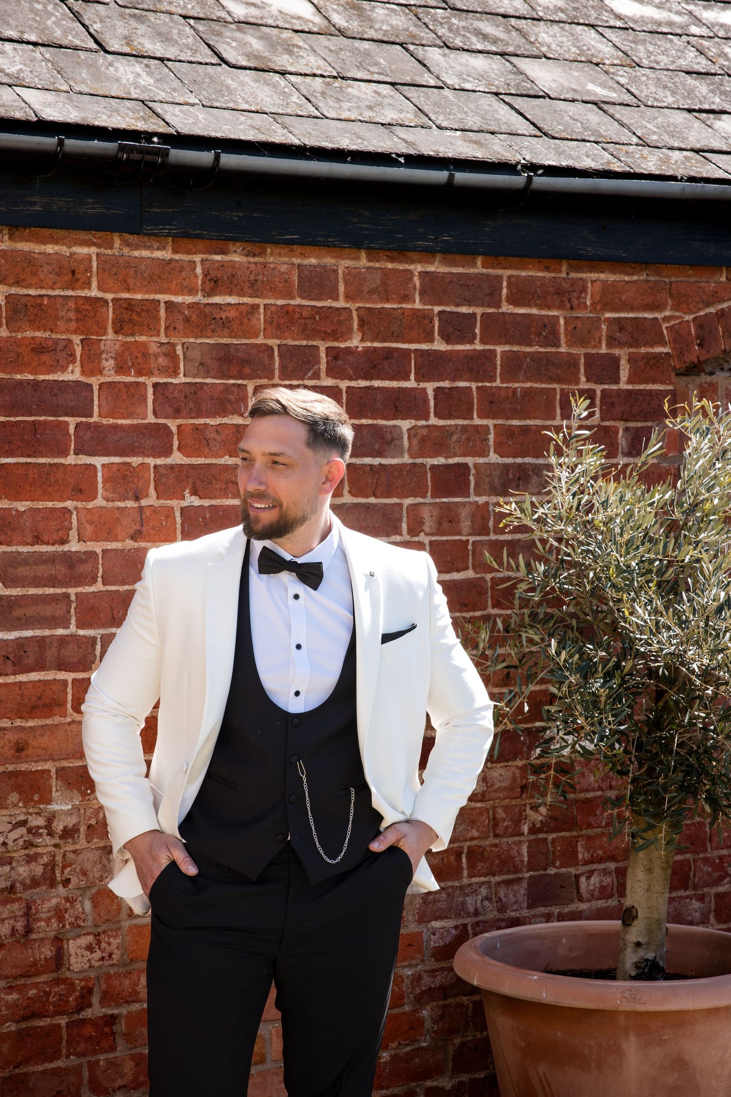Groom in white tuxedo jacket and bow tie posing outdoors against a red brick wall with a potted olive tree.