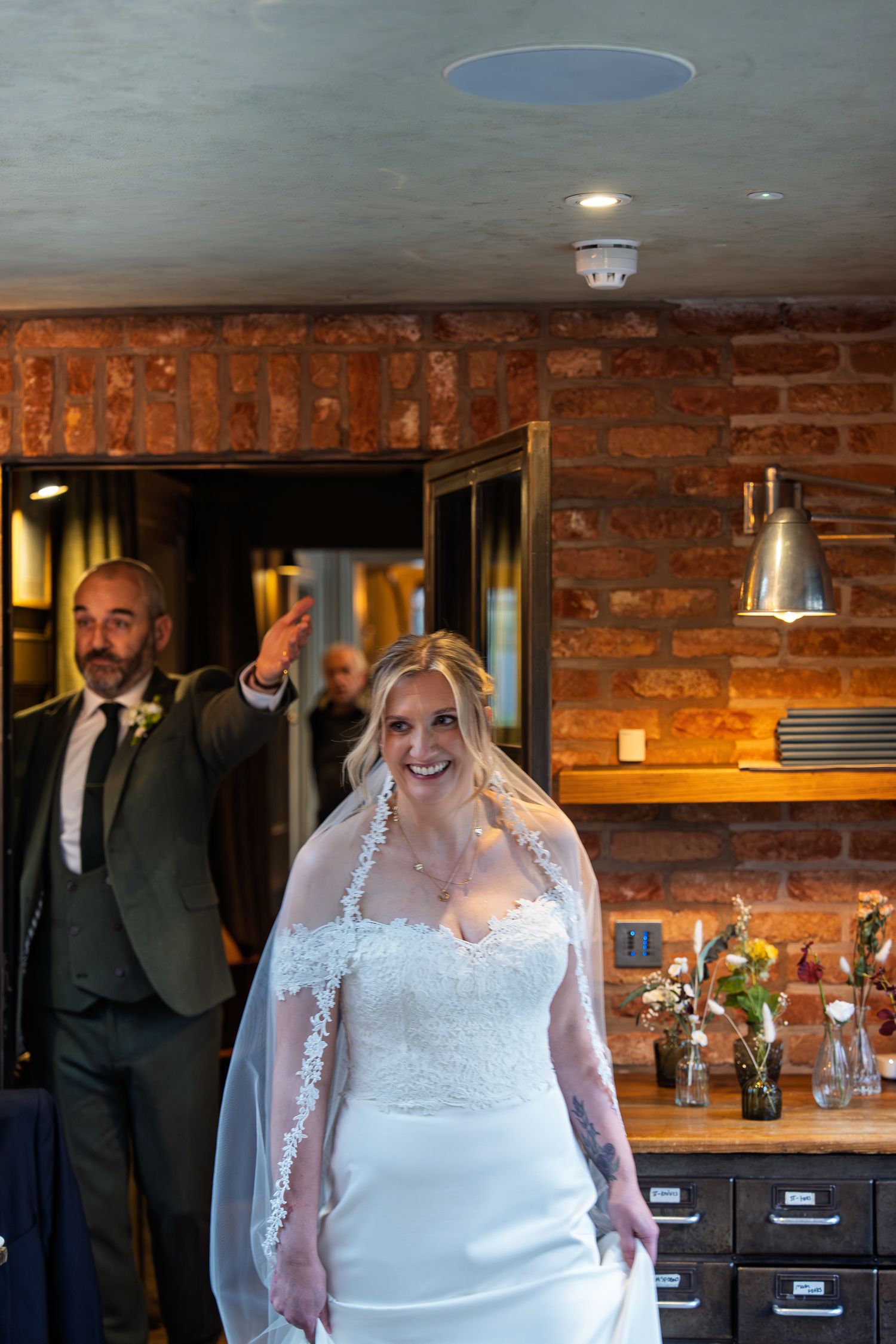 Smiling bride in lace wedding dress and veil enters rustic brick venue as groom waves behind her.