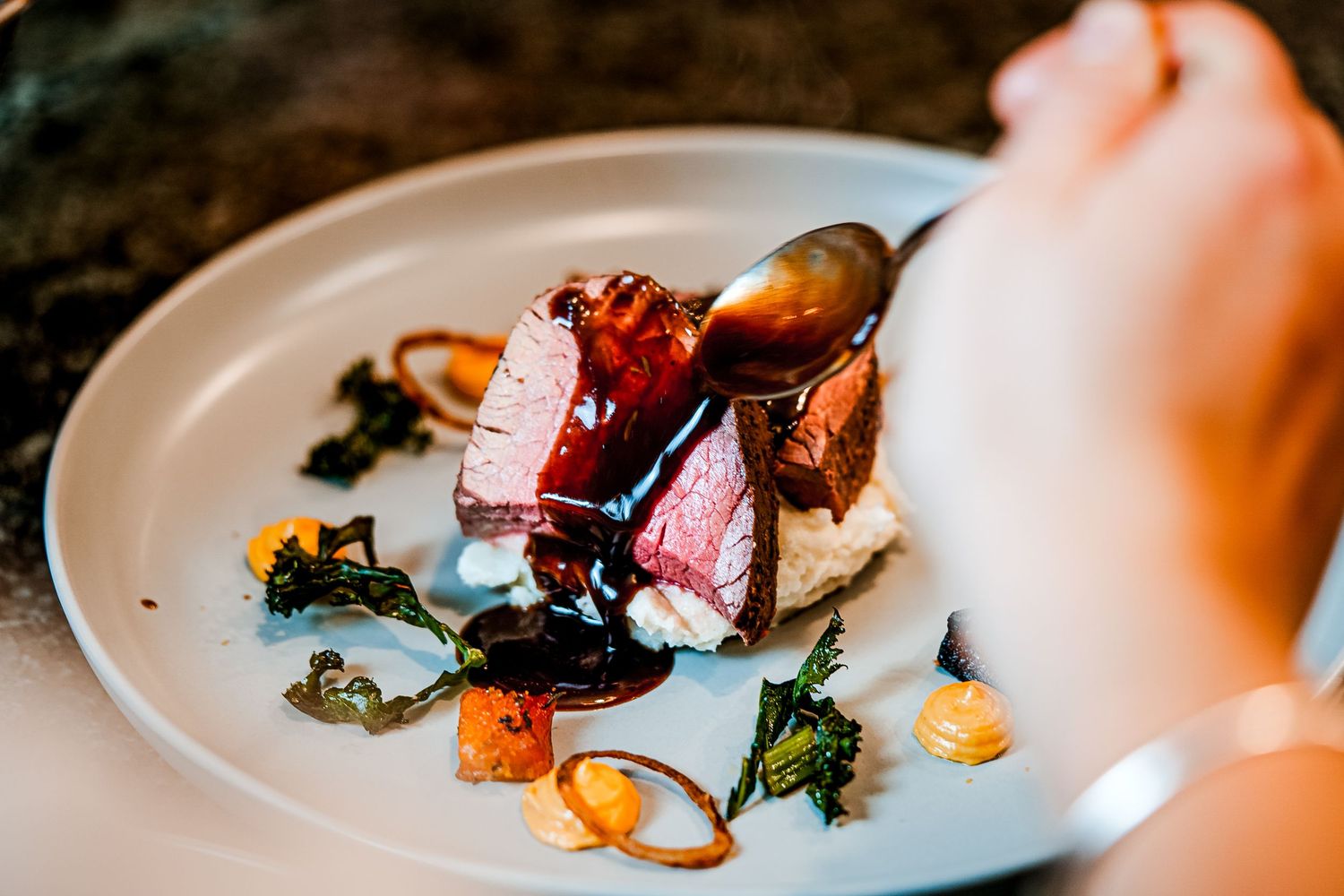Gourmet plated dish with sliced medium-rare meat, garnished with greens, sauce, and colorful vegetables on a white plate.