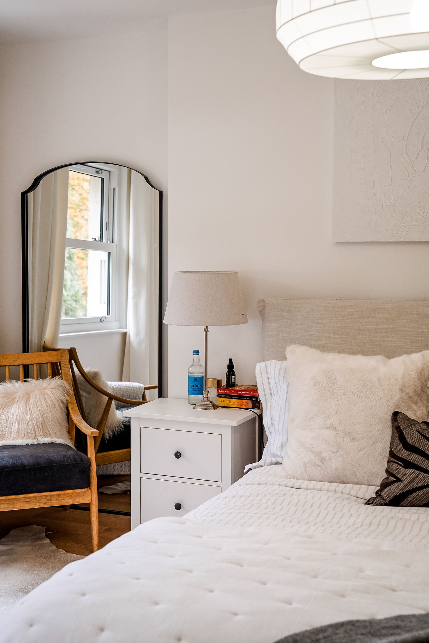 Bright bedroom with white bedding, nightstand, floor mirror, wooden chair, and pendant light in a cozy neutral space.