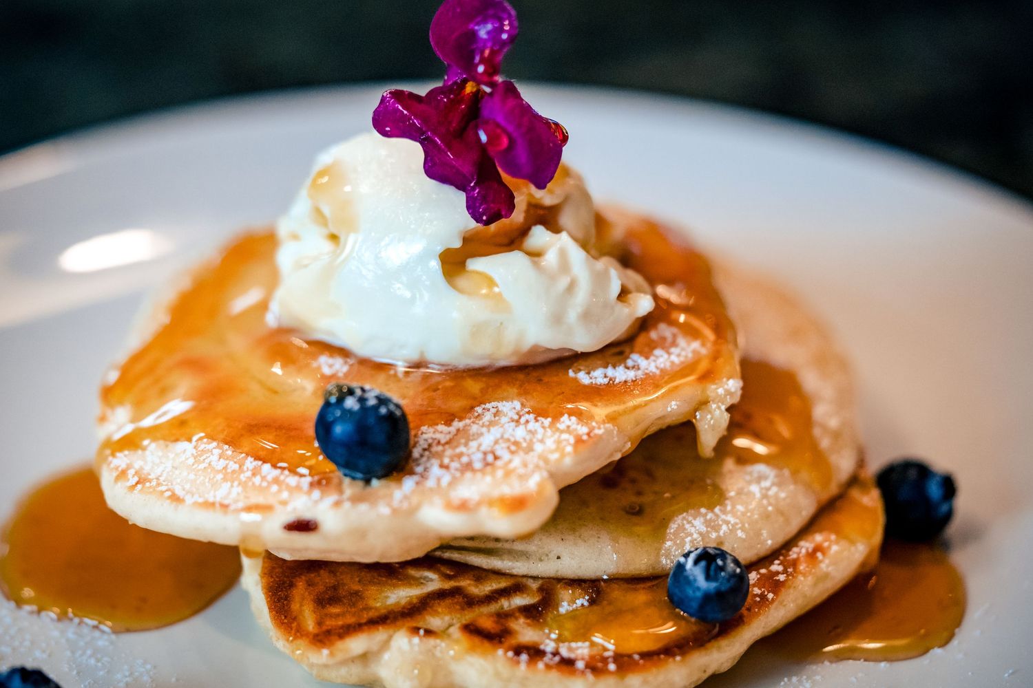 Stack of fluffy pancakes topped with whipped cream, blueberries, syrup, and a purple orchid on a white plate.