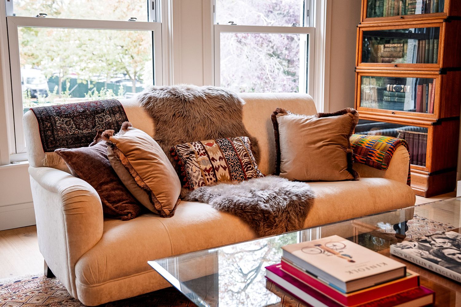 Cozy beige sofa with decorative pillows and fur throw near a bright window, with a glass coffee table and bookcase.