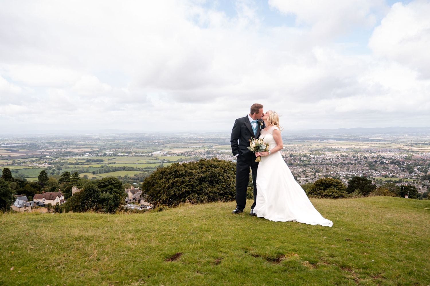 Bride and groom share a romantic kiss on a hilltop with a sweeping countryside panorama behind them.