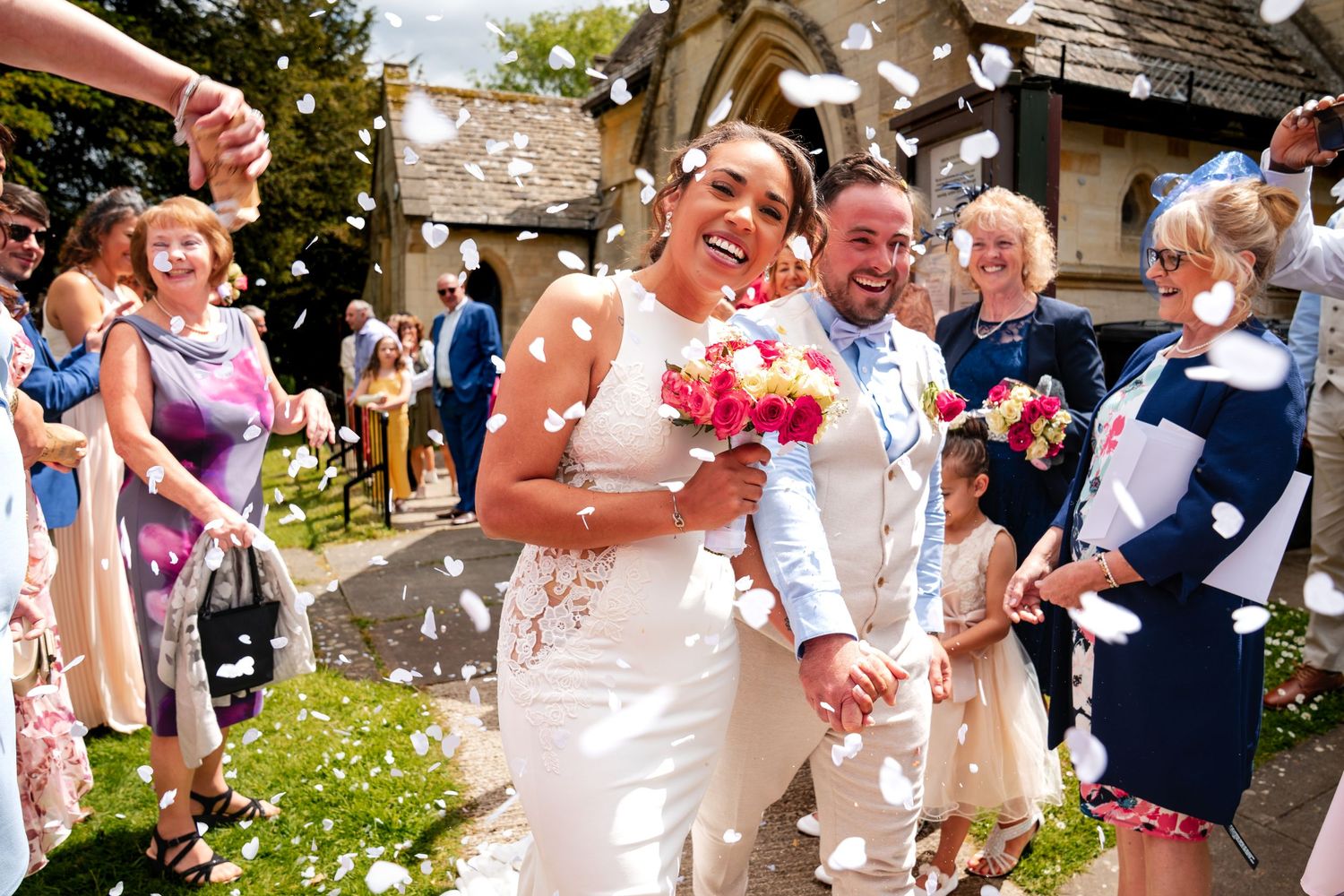 Newlywed couple walking through confetti shower outside church, bride holding pink bouquet, guests celebrating wedding ceremony.