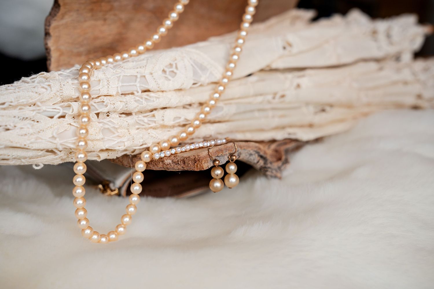 Elegant pearl necklace and earrings draped over rustic driftwood on white fur, vintage jewelry display.