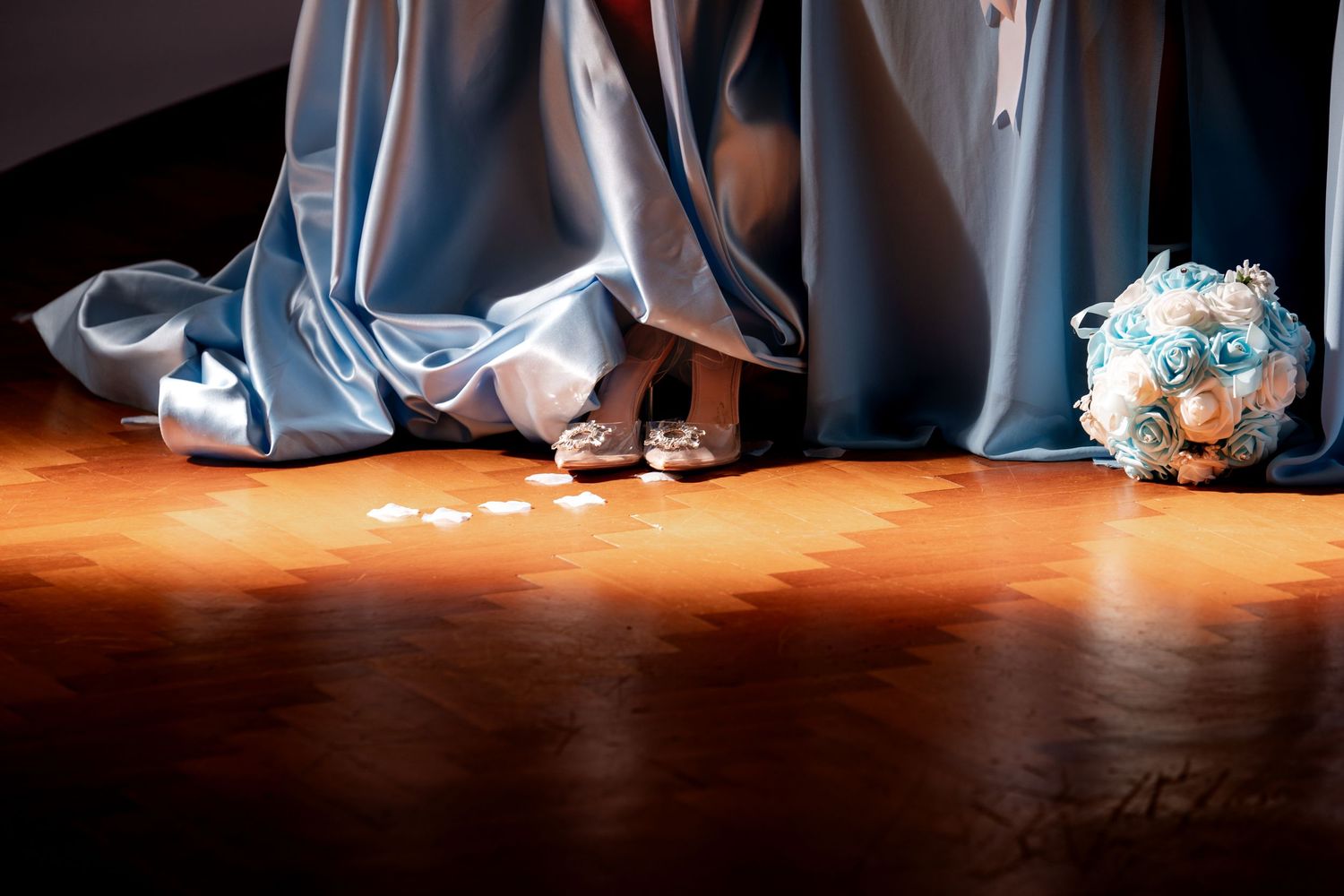 Bride in blue gown with silver heels stands near a blue rose bouquet on a wooden floor.