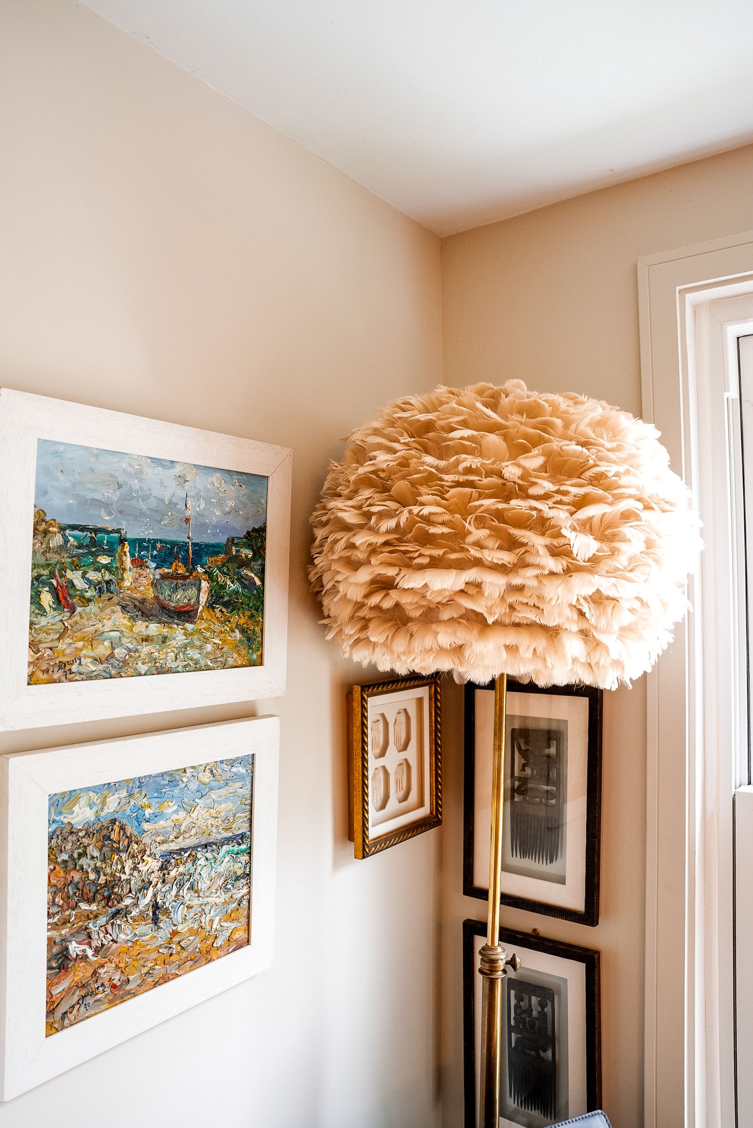A cozy corner with a fluffy feather pendant lamp, framed coastal paintings, and gallery wall art near a white door.