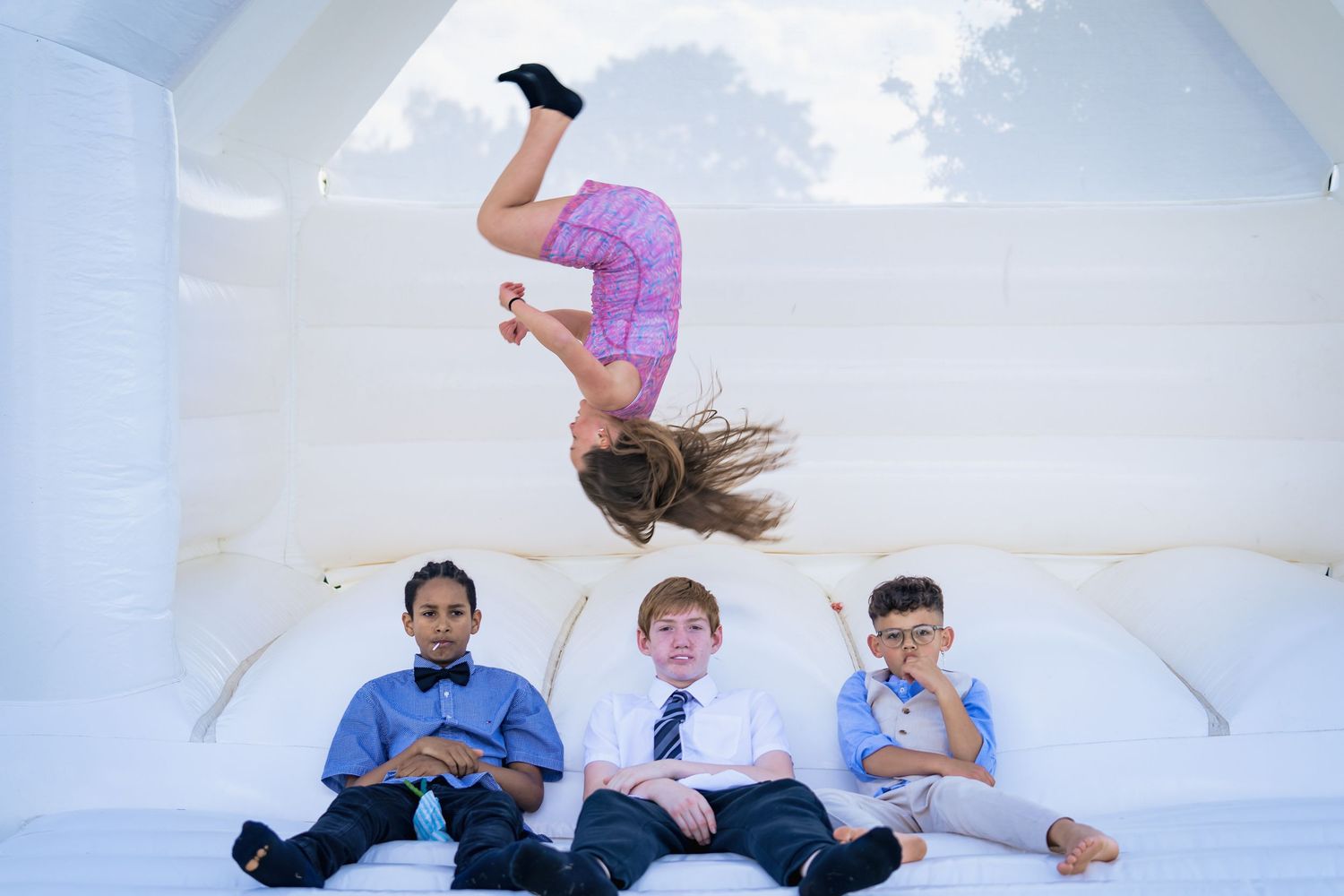 Girl doing backflip on white bouncy castle while three boys in school uniforms sit watching below.