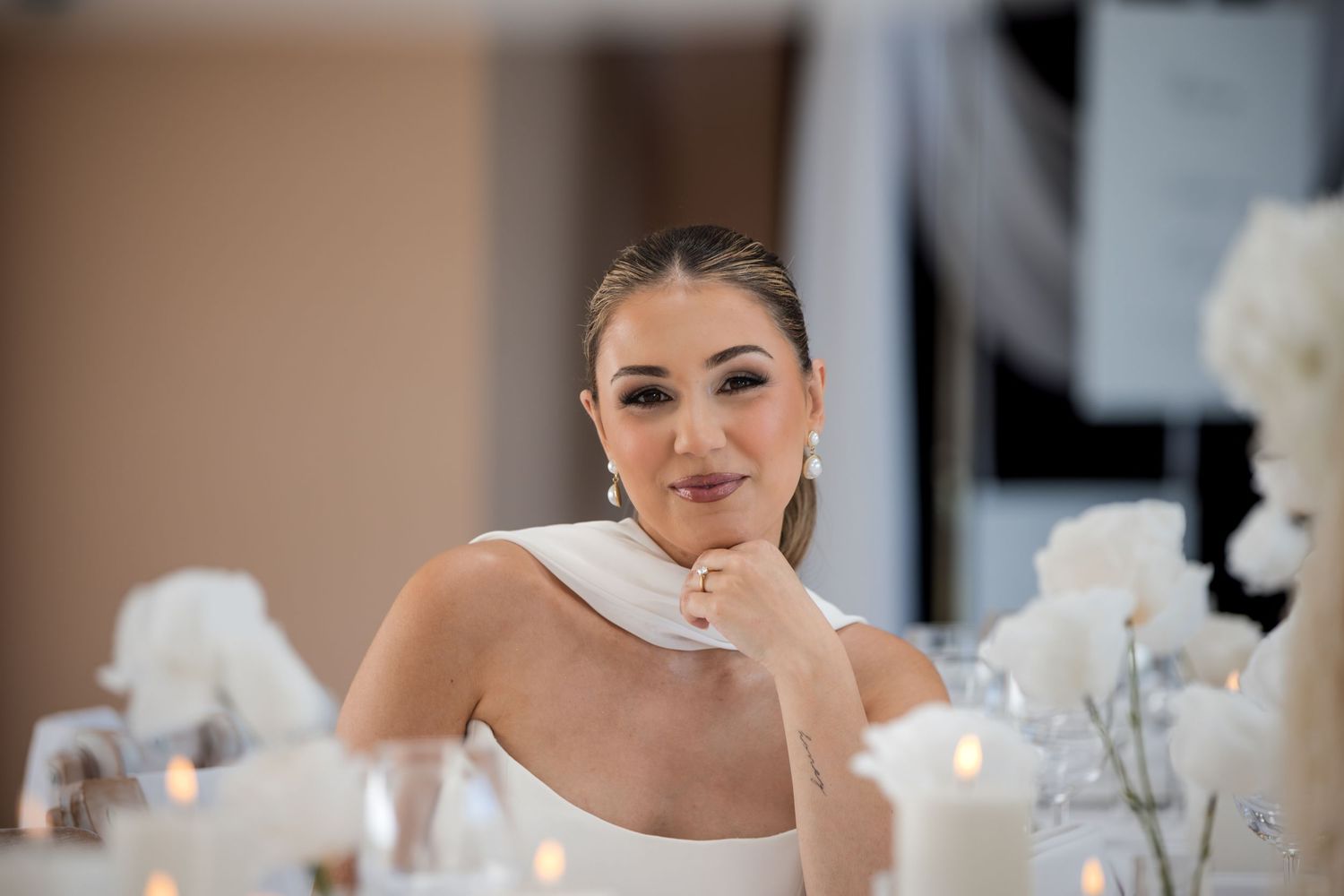 Elegant bride in white dress sits at reception table with candles and floral arrangements, resting chin on hand.