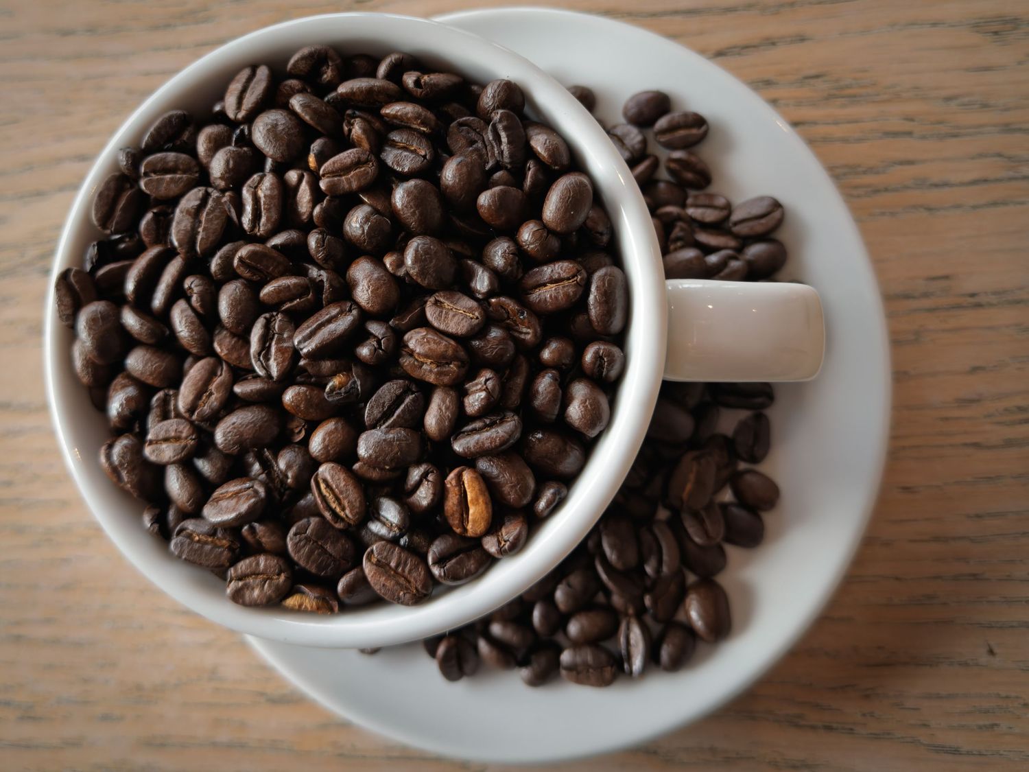 A white ceramic cup and saucer overflowing with roasted coffee beans on a wooden surface.