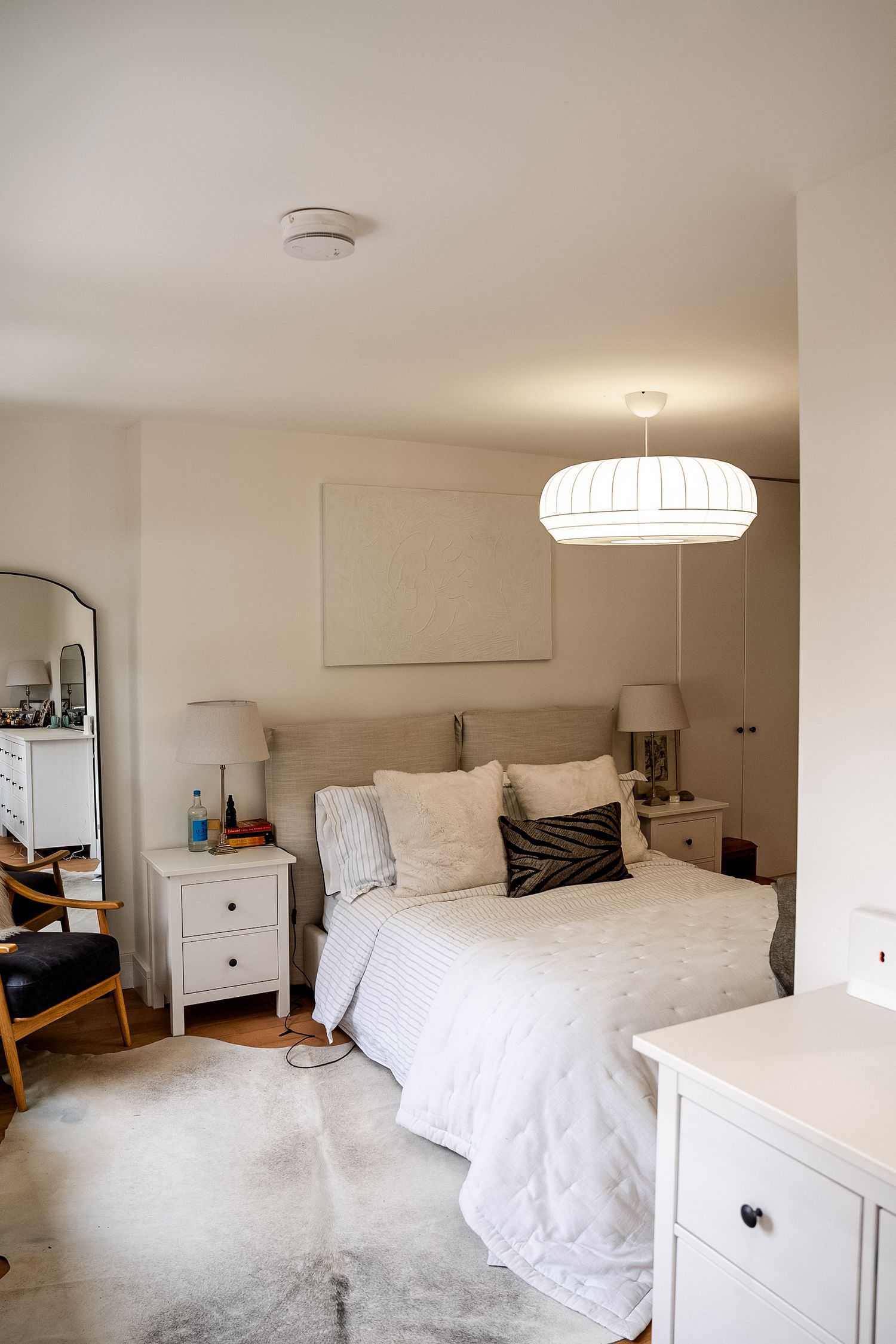 Bright white bedroom with cozy bedding, pendant light, nightstands, fur rug, and a large mirror leaning against the wall.
