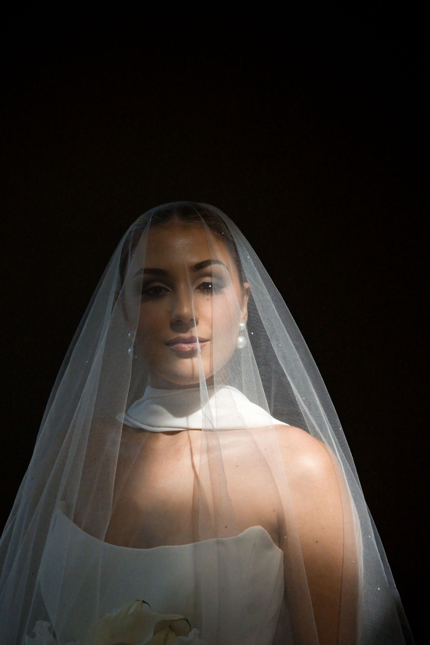 Elegant bride wearing a sheer white veil over her face, posing against a dark background in a wedding dress.