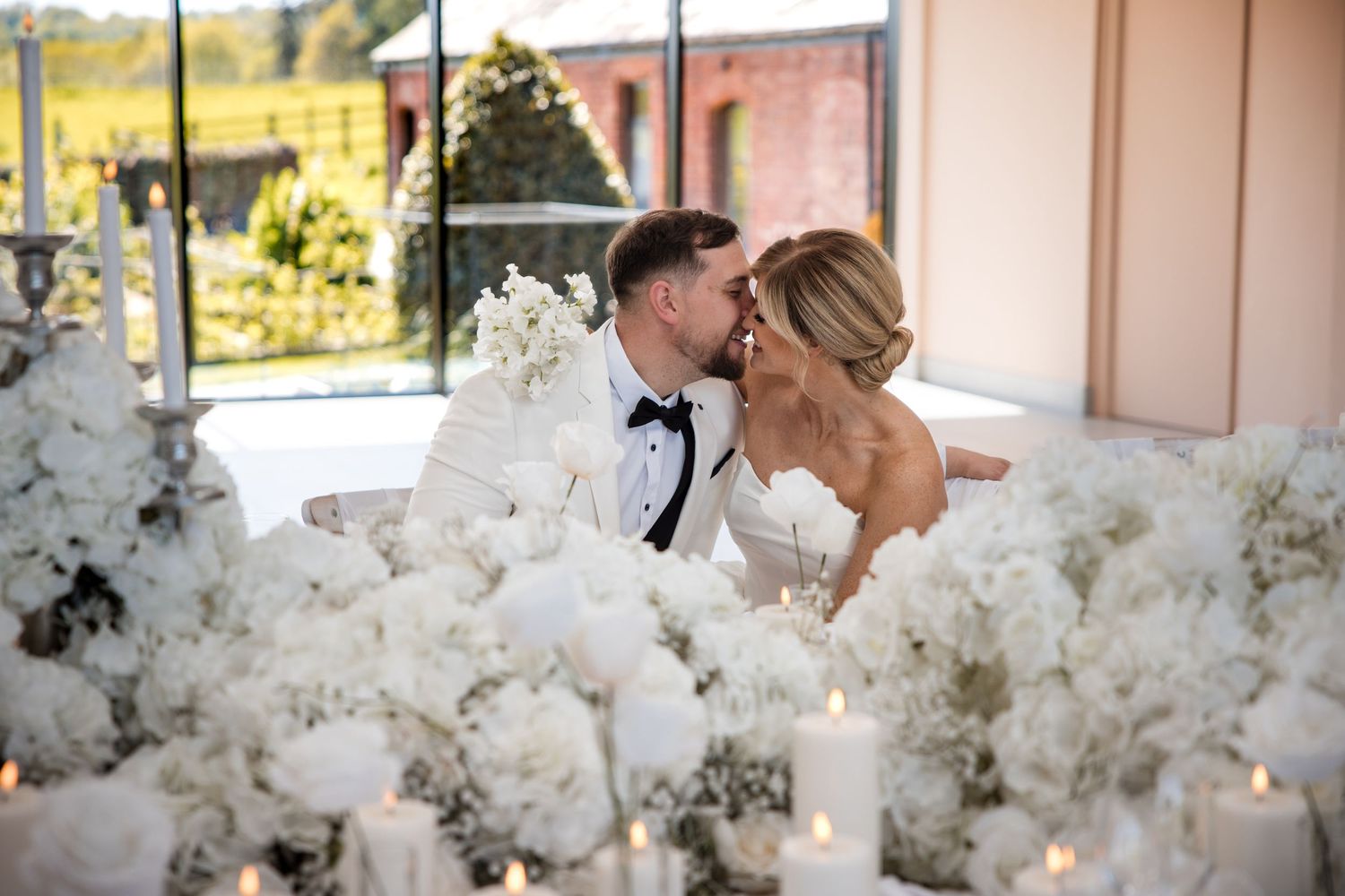 Bride and groom share a romantic kiss surrounded by white floral arrangements and glowing candles at their elegant wedding reception.