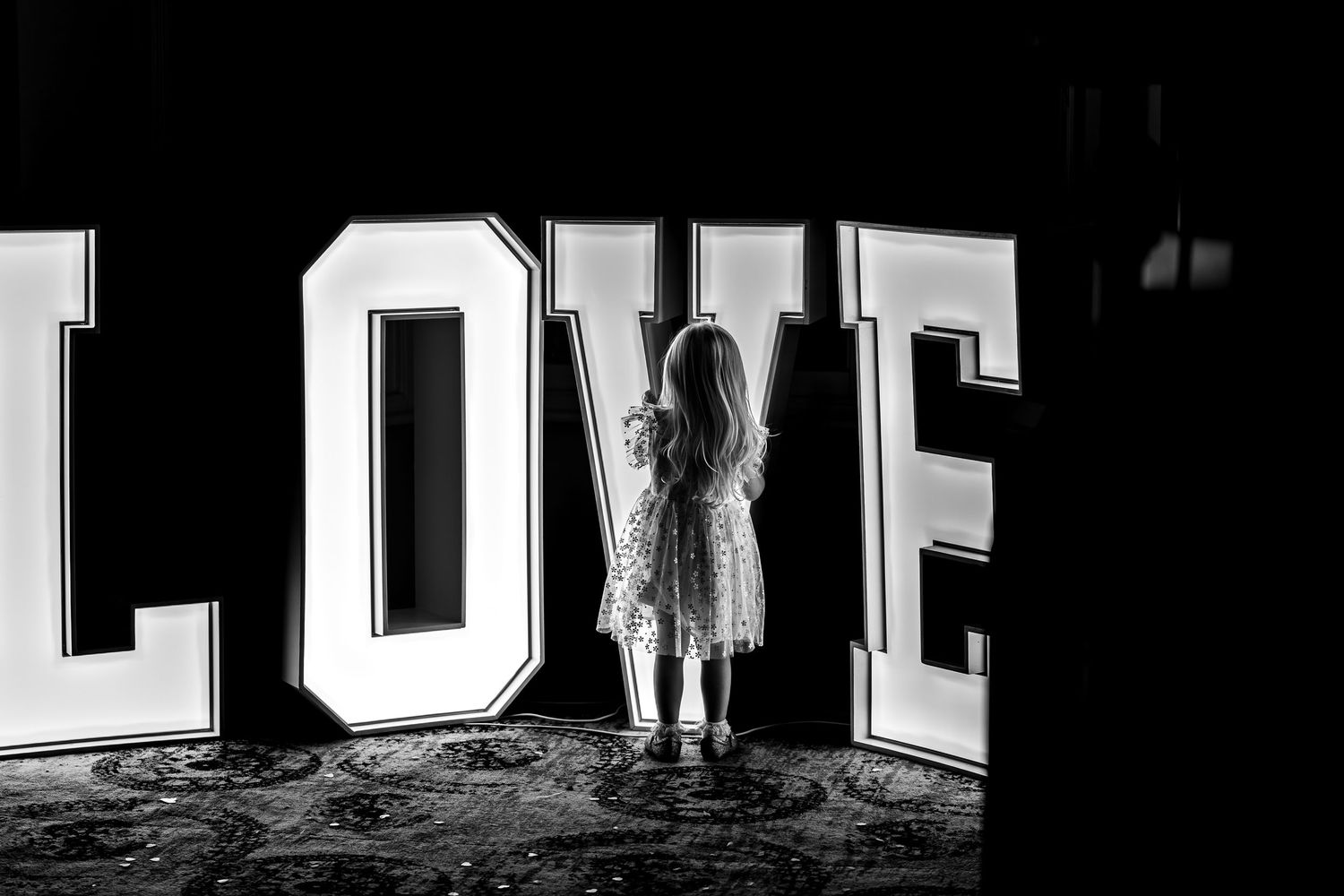 Black and white photo of a young girl standing before illuminated LOVE letters at an event.