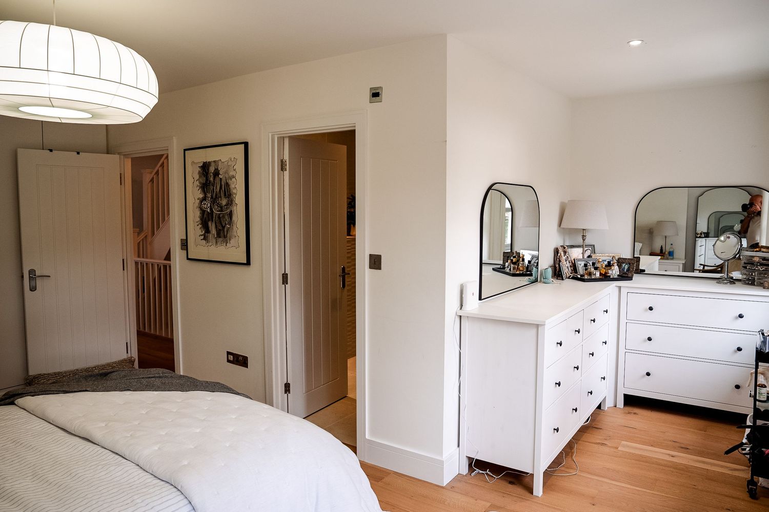 Bright bedroom with white dresser, arched mirrors, pendant light, hardwood floors, and open door revealing hallway.