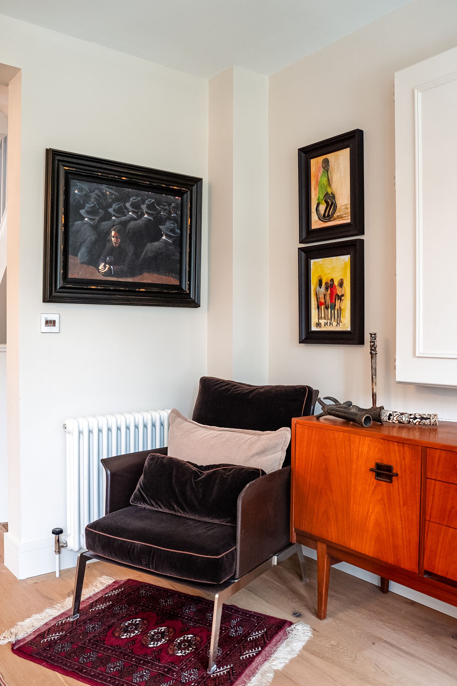 Cozy reading corner with dark velvet armchair, mid-century sideboard, red patterned rug, and colorful framed artwork on white walls.