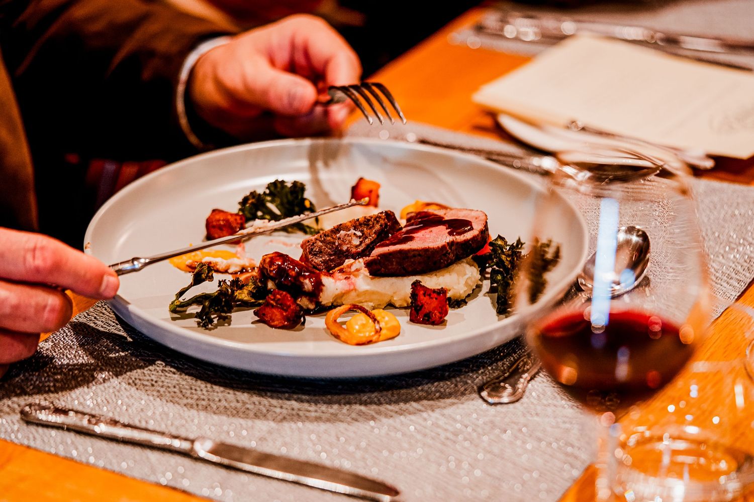 Person cutting into a gourmet meat dish with roasted vegetables on a white plate, with a glass of red wine nearby.