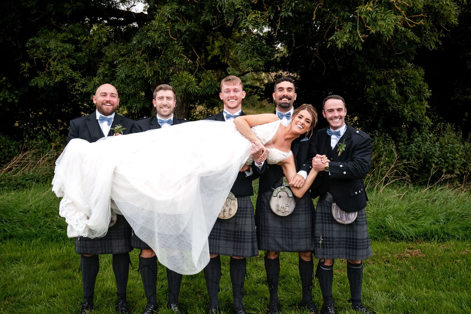 Groom and groomsmen in Scottish kilts lifting the bride outdoors at a wedding celebration.