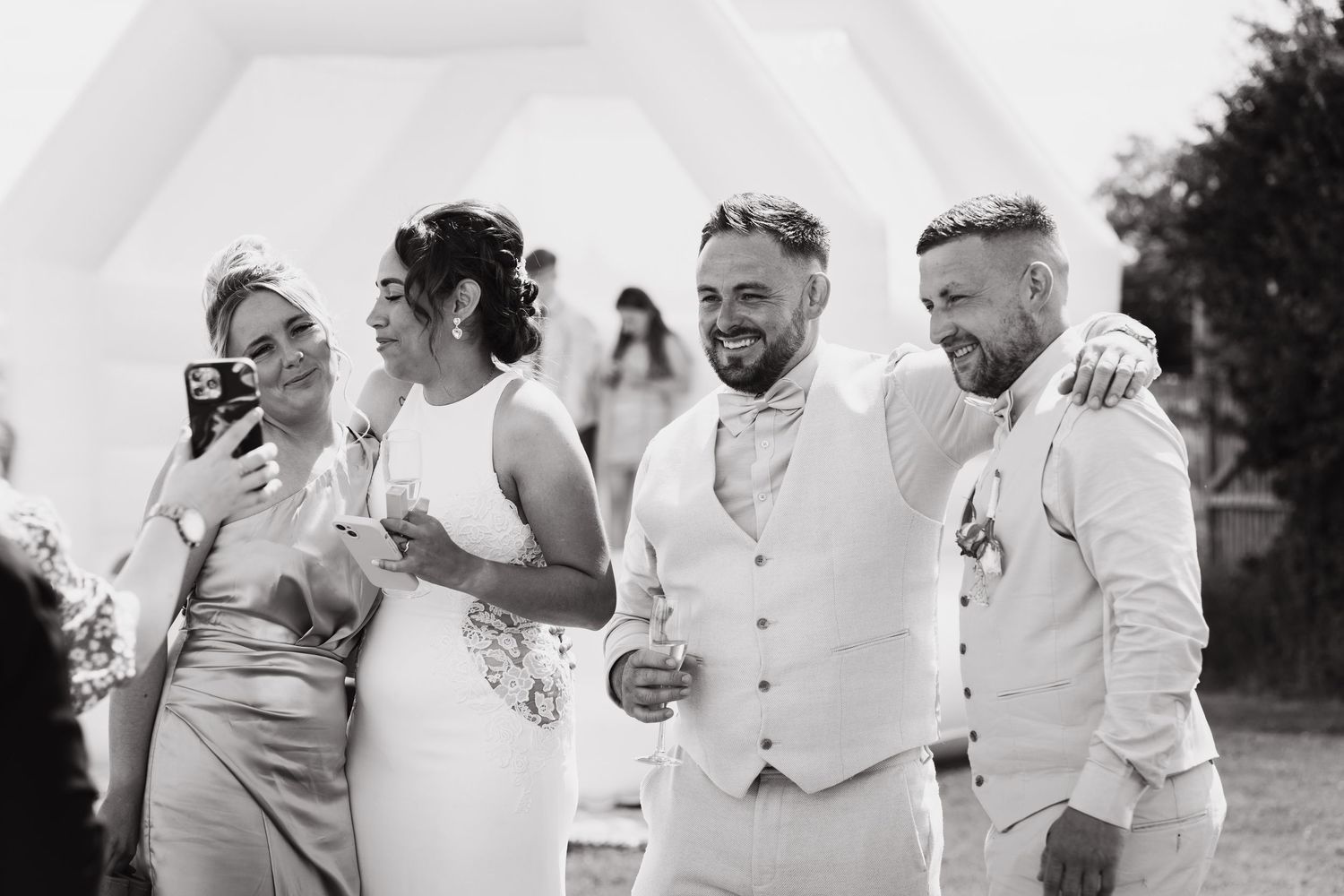 Black and white wedding photo of bride, groom, and guests laughing outdoors at a sunny celebration.