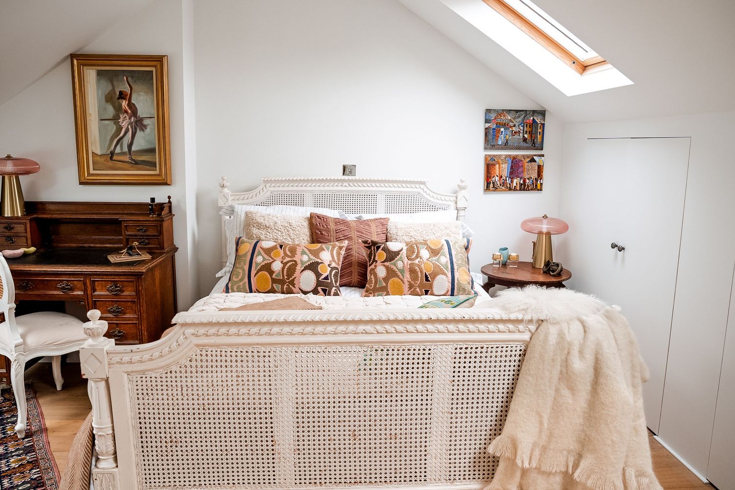 Cozy attic bedroom with white cane bed, floral pillows, skylight, vintage dresser, and warm pink lamp accents.